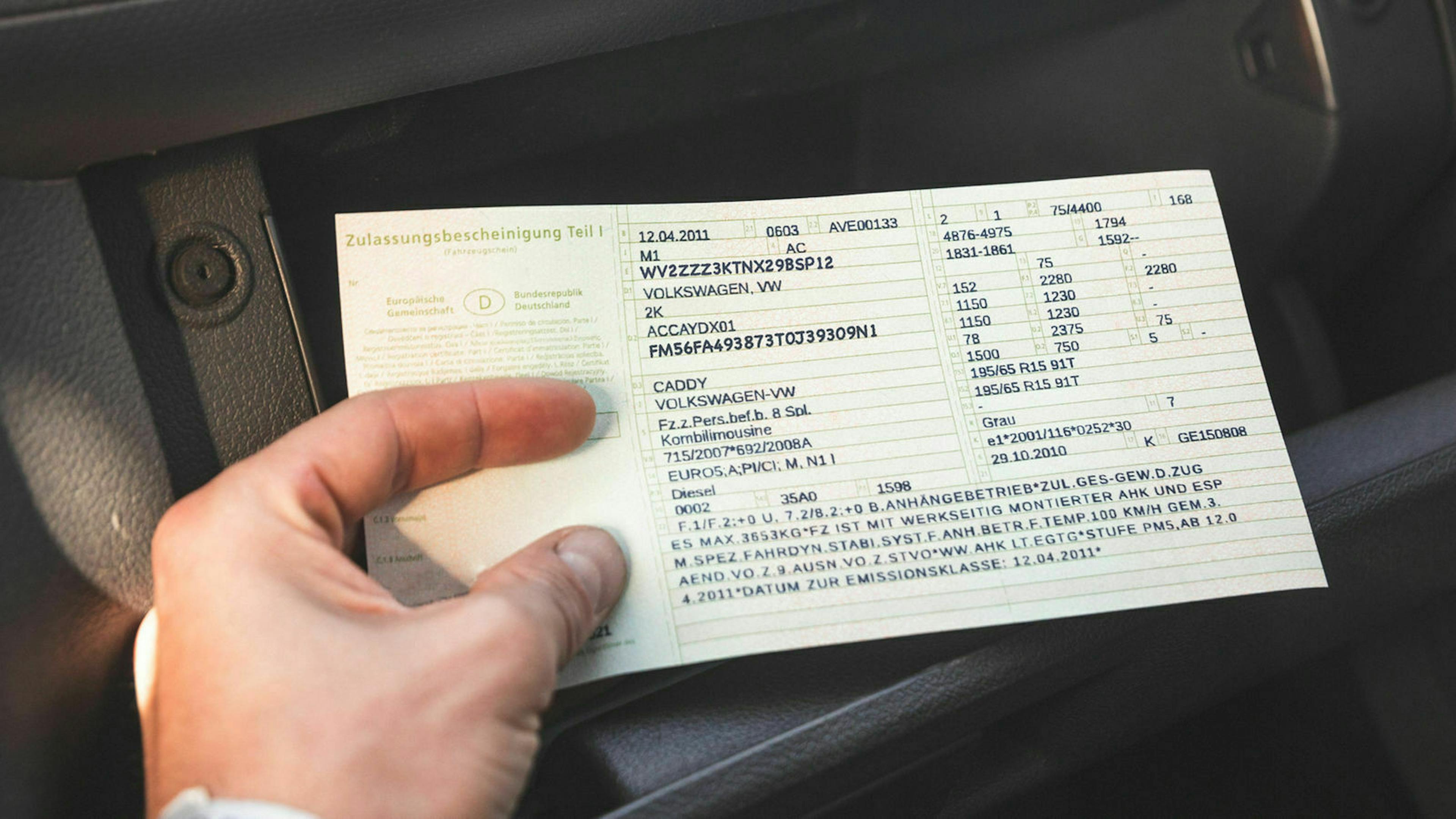 Jemand hält einen Fahrzeugschein in der Hand. Im Hintergrund befindet sich das Handschuhfach eines Autos