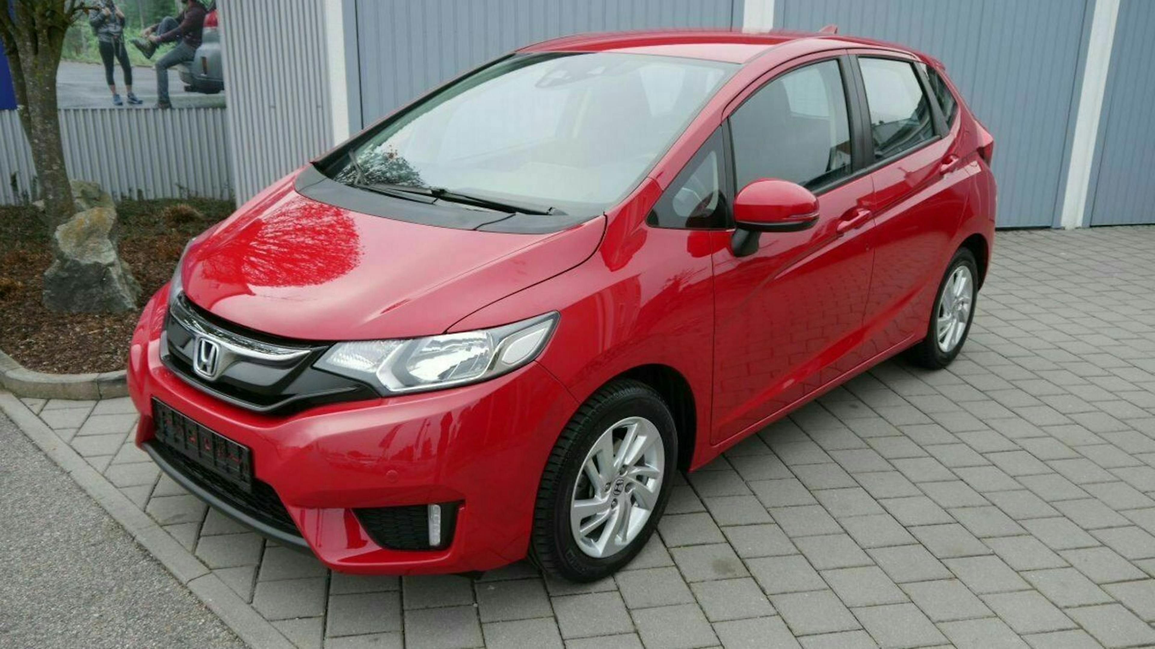 Roter Honda Kleinwagen auf gepflasterter Fläche, geparkte Autos im Hintergrund, moderner Stil mit glänzendem Lack.