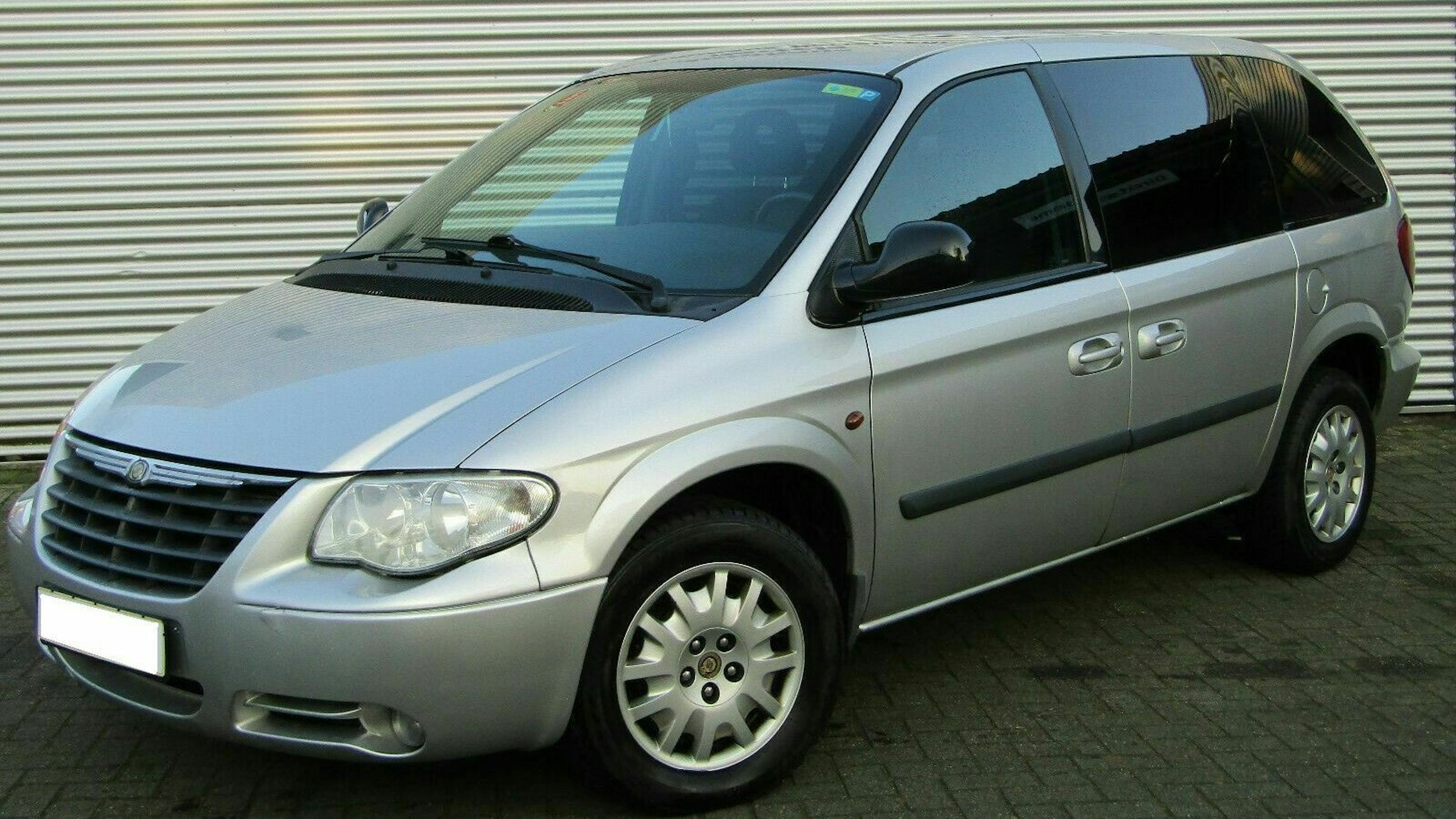 Silberner Chrysler Voyager Minivan vor einem weiß-geriffelten Hintergrund, seitliche Ansicht, geparkt auf einem gepflasterten Untergrund.