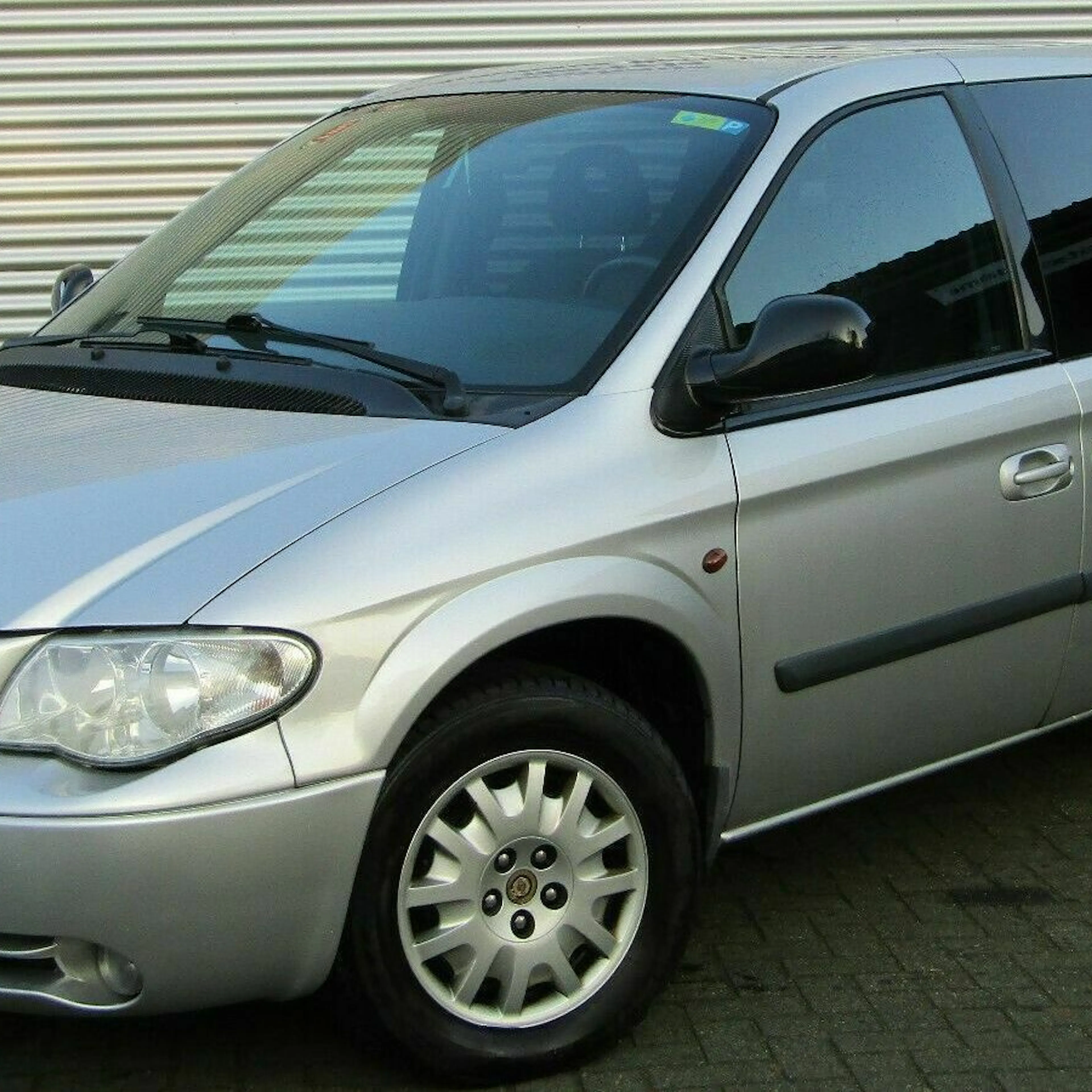 Silberner Chrysler Voyager Minivan vor einem weiß-geriffelten Hintergrund, seitliche Ansicht, geparkt auf einem gepflasterten Untergrund.