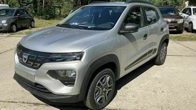 Ein silberner Dacia Spring steht auf einem Parkplatz, im Hintergrund sind Bäume und weitere PKWs zu erkennen