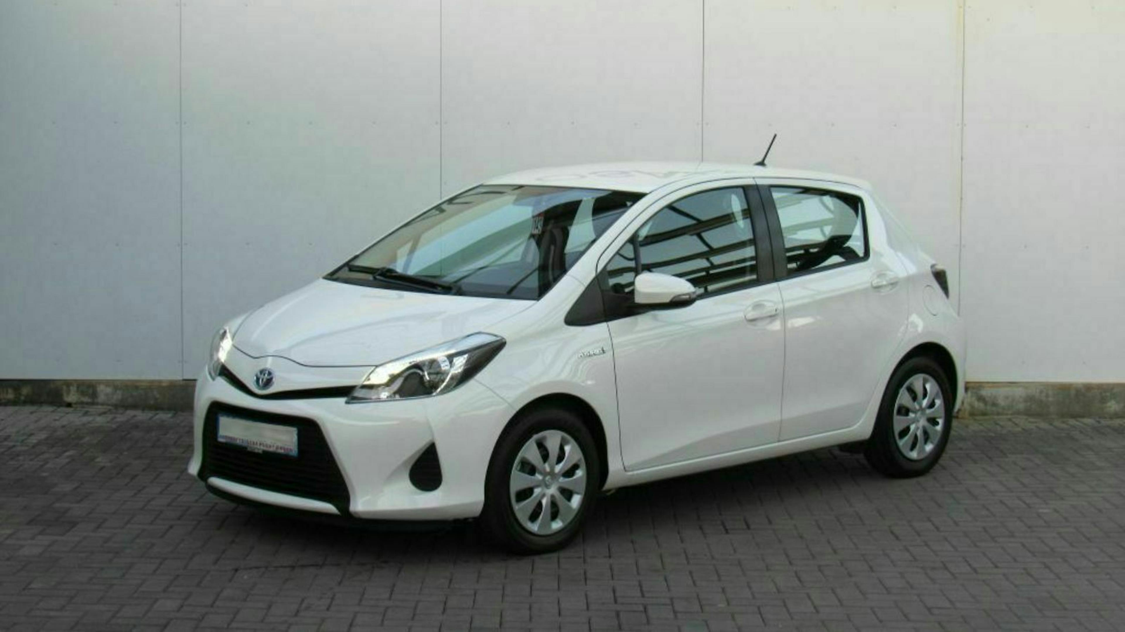 Weißer Kleinwagen, geparkt vor einer grauen Wand auf gepflastertem Boden, mit deutlich sichtbarer Front und Seite.