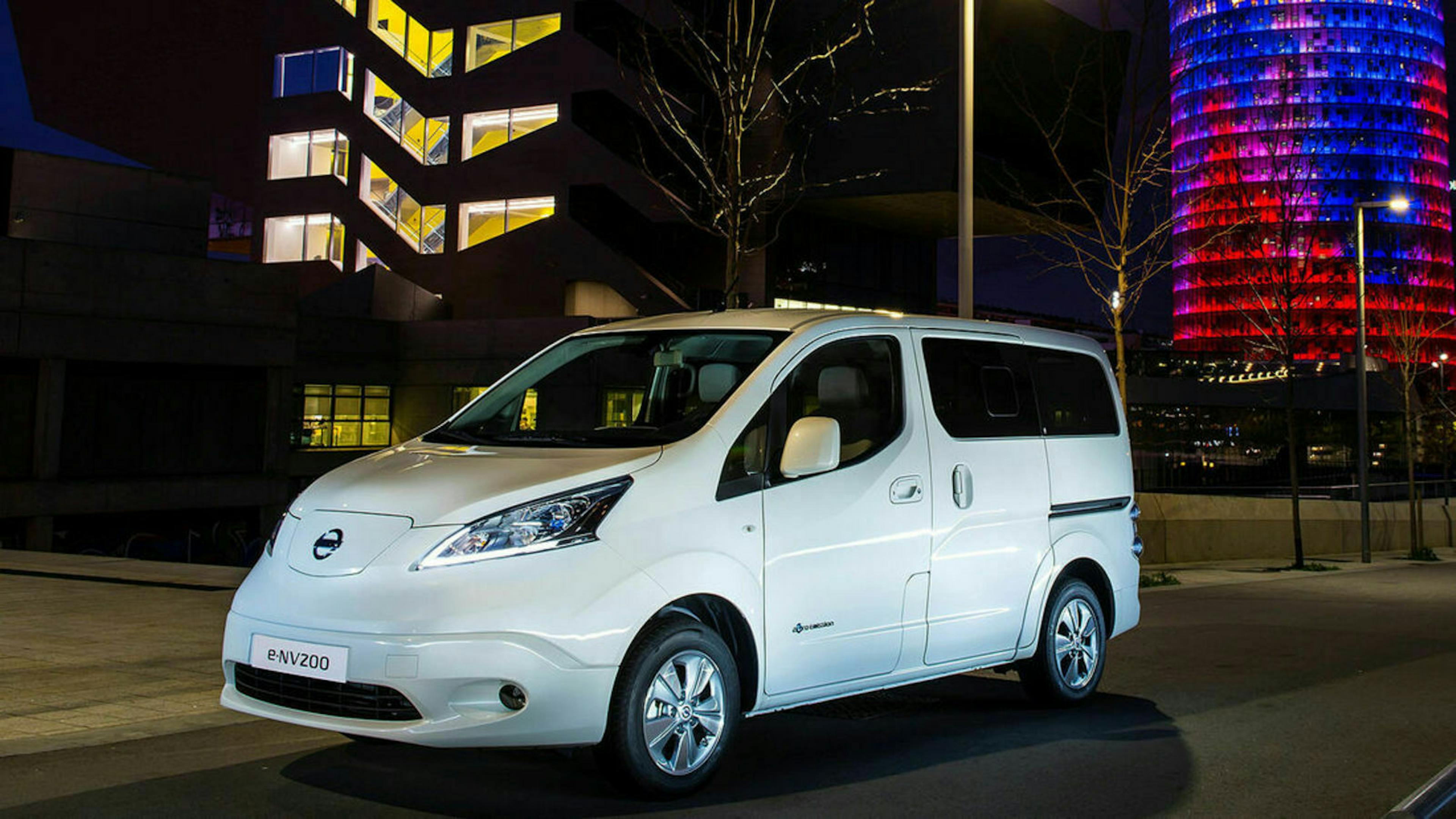 Ein weißer Nissan eNV200 steht bei Nacht vor einem Bürogebäude, in dem einige Lichter eingeschaltet sind