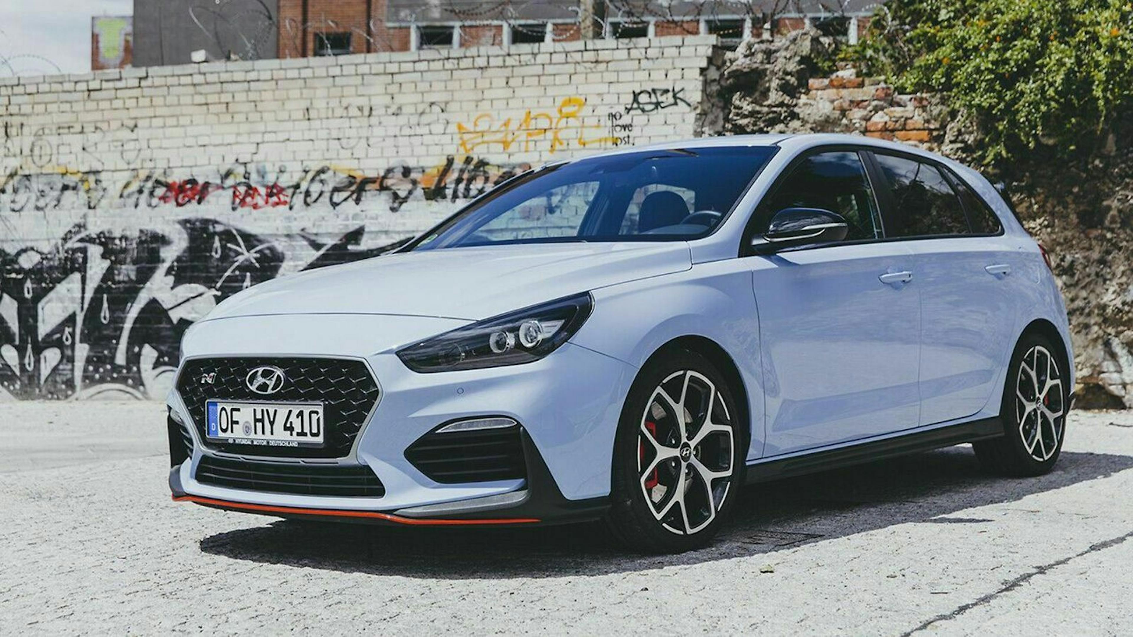 Weißer Hyundai i30N vor einer grauen Mauer mit Graffiti, geparkter Kompaktwagen auf hellem Pflaster.