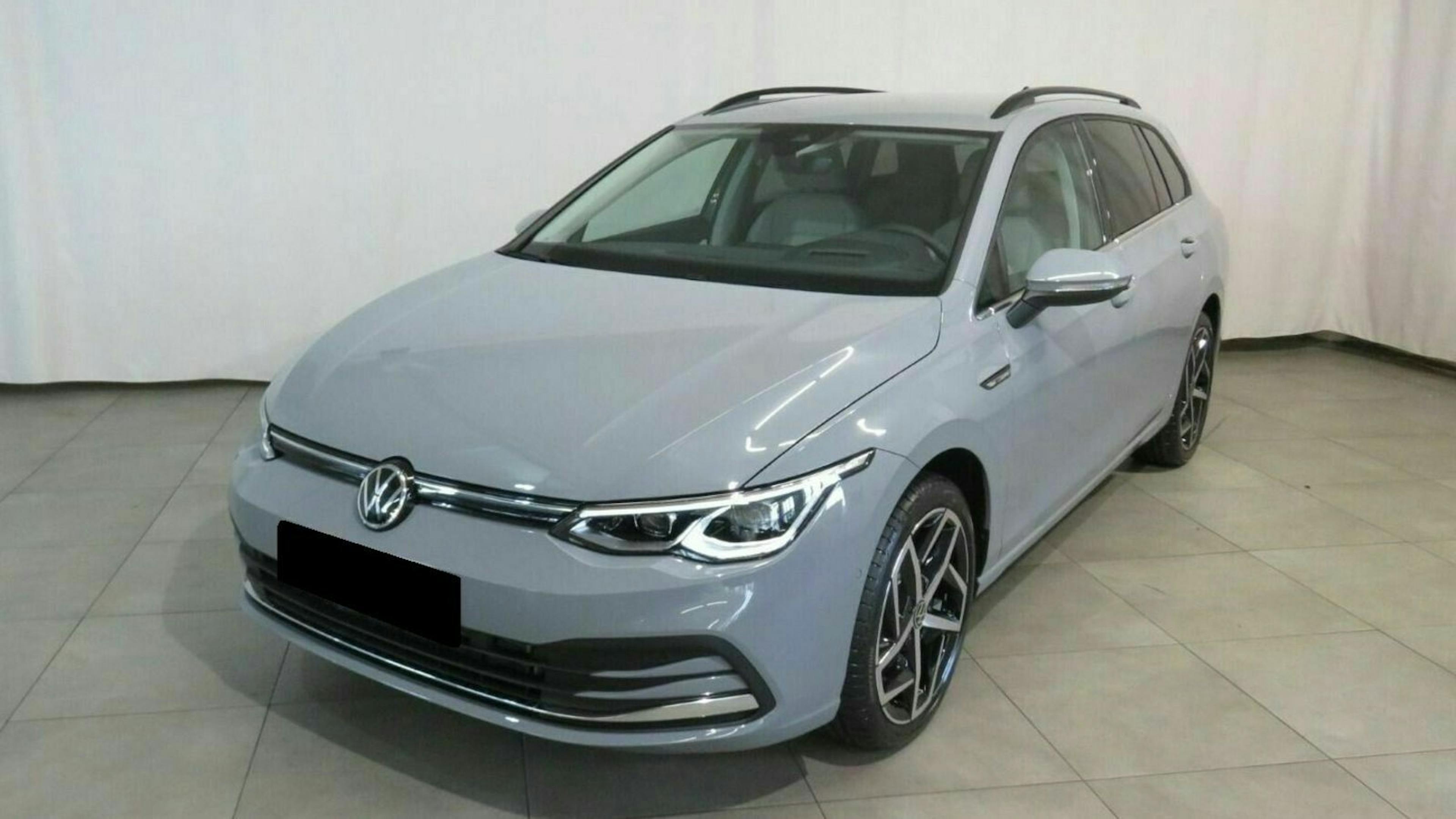 Silberner VW Kombi steht in einem Ausstellungsraum mit Fliesenboden und weißem Hintergrund. Fahrzeug zeigt Front- und Seitenansicht.