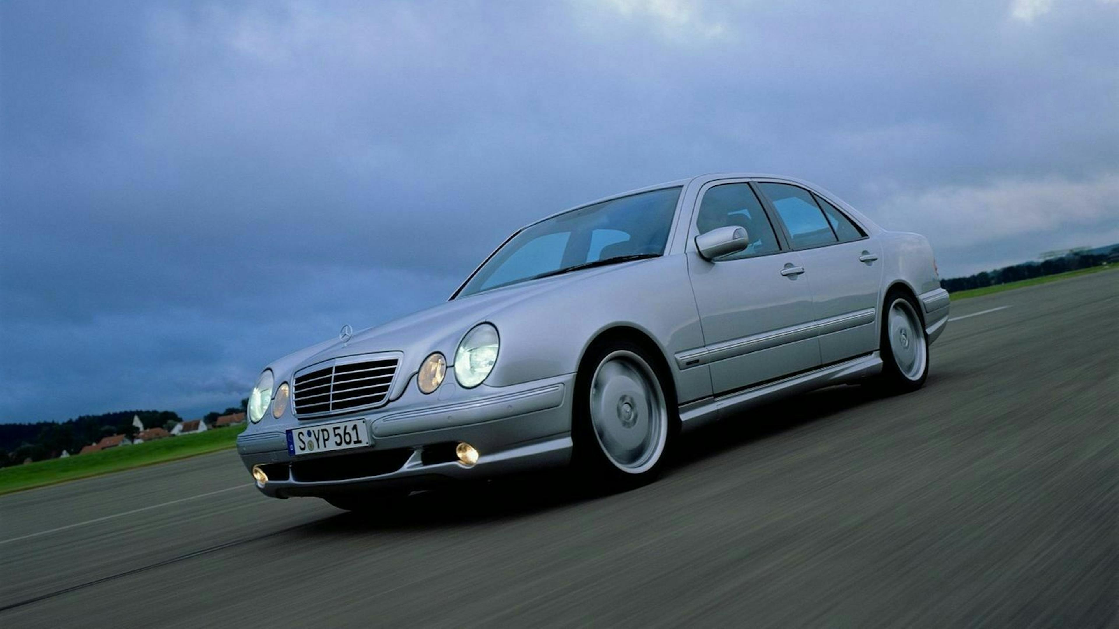 Ein silberner Mercedes-Benz E55 AMG W210 Oldtimer fährt eine Landstraße entlang.