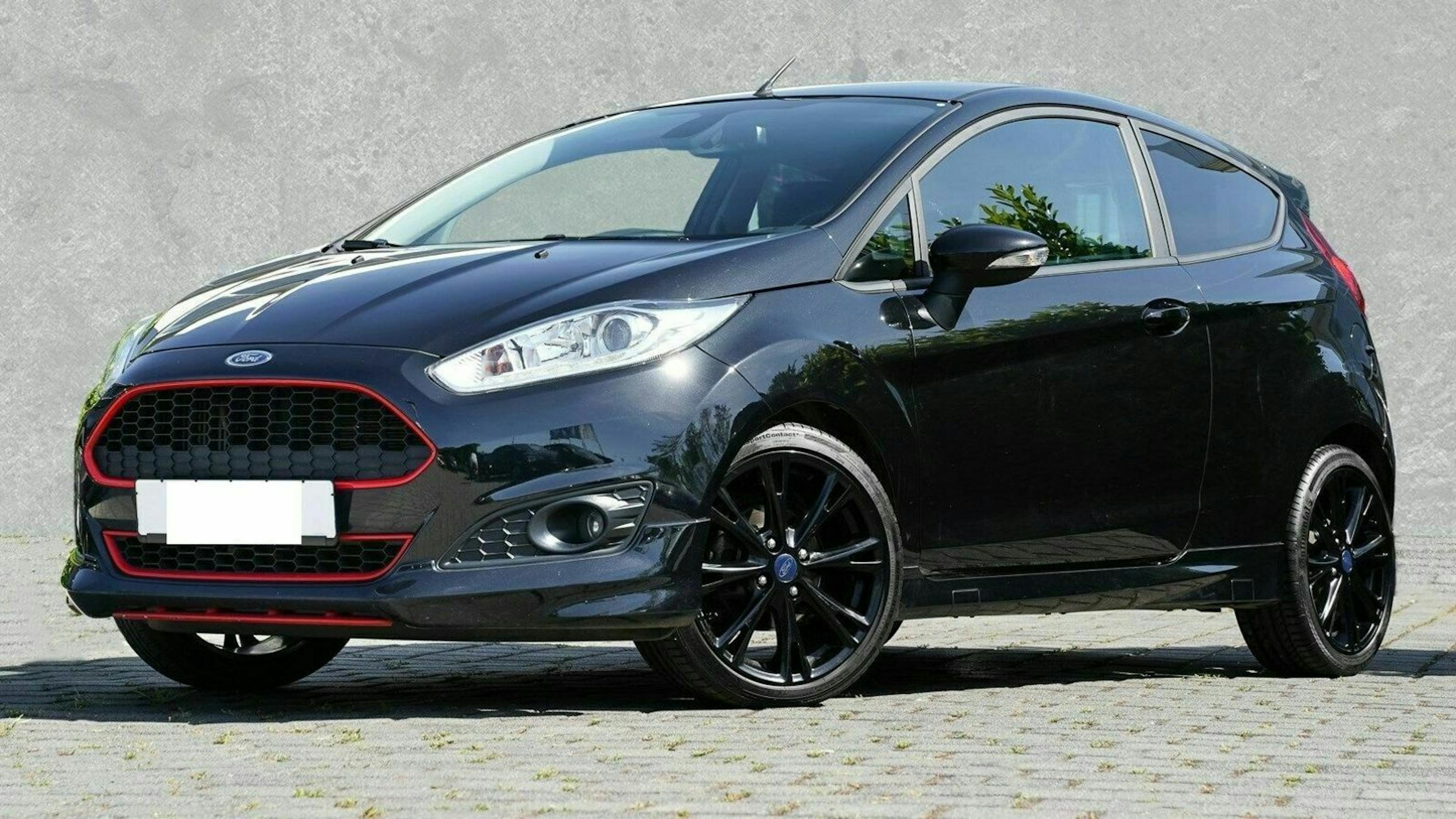 Schwarzer Ford Fiesta mit roten Akzenten, auf einem gepflasterten Platz geparkt, grauer Hintergrund.