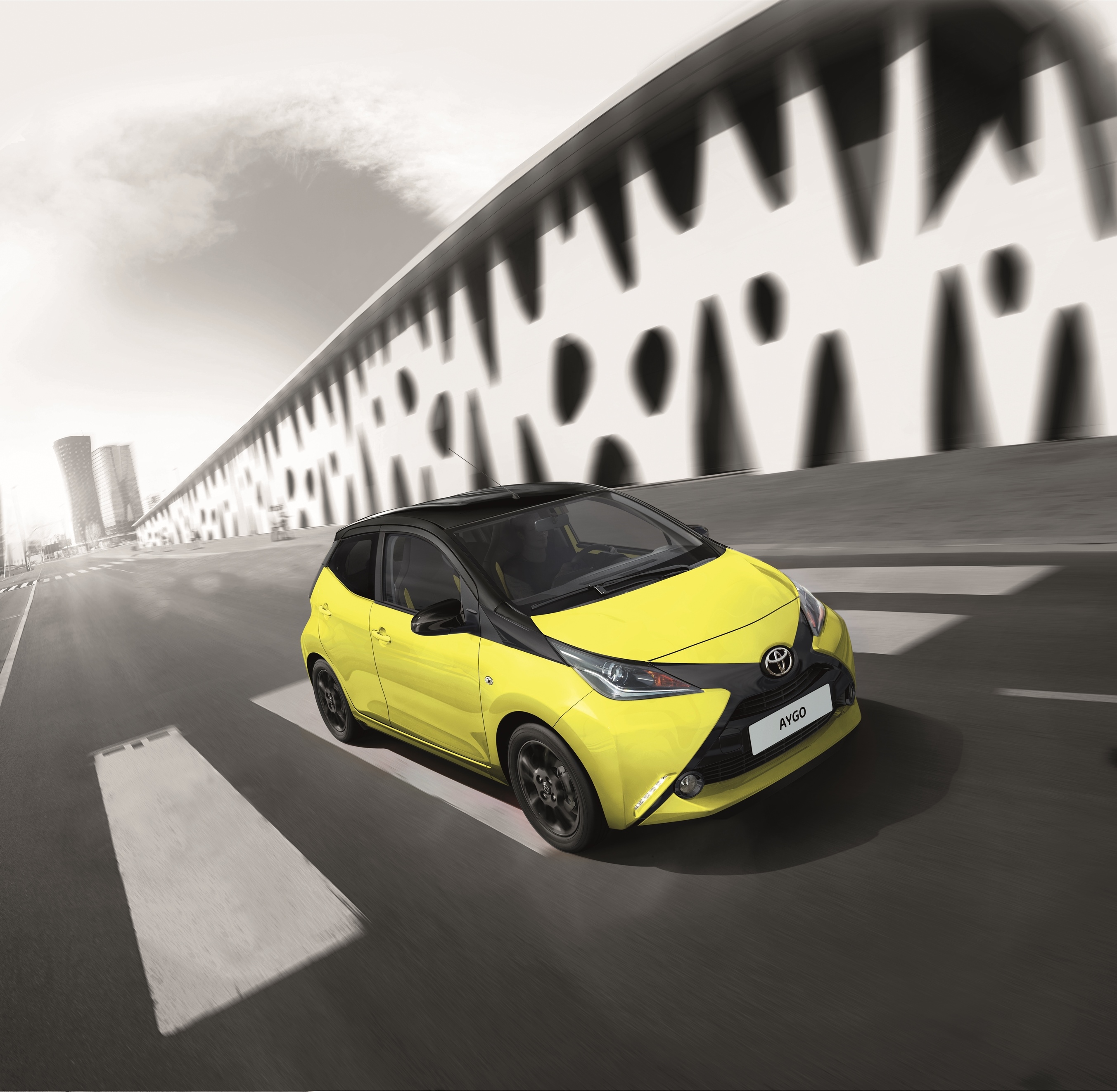 Gelber Toyota Aygo mit schwarzem Dach und schwarzem X-Element in der Front fährt vor einer weißen Wand mit großem Muster.