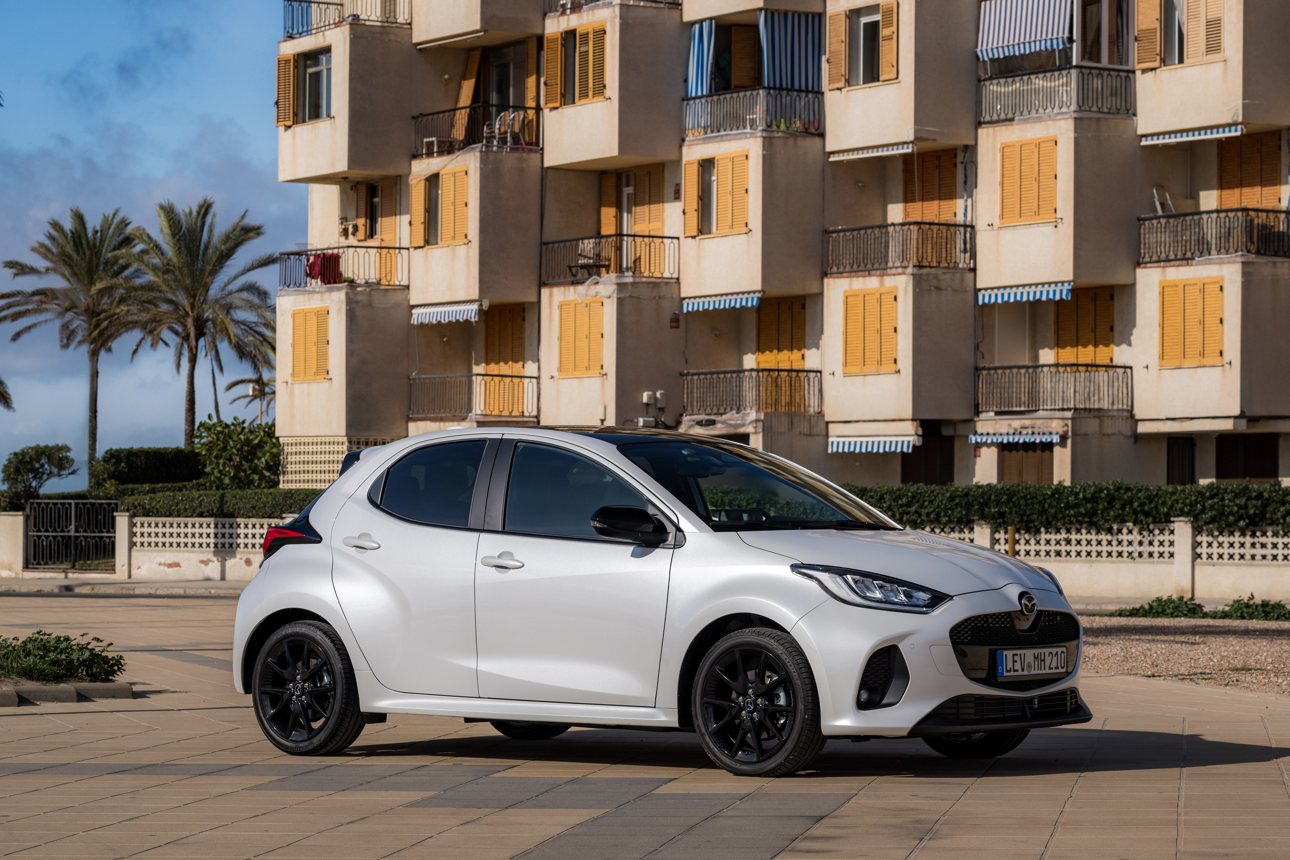 Weißer Mazda2 Hybrid mit schwarzem Dach und schwarzen Leichtmetallfelgen vor einem Wohnhaus mit gelben Fensterläden und Palmen.
