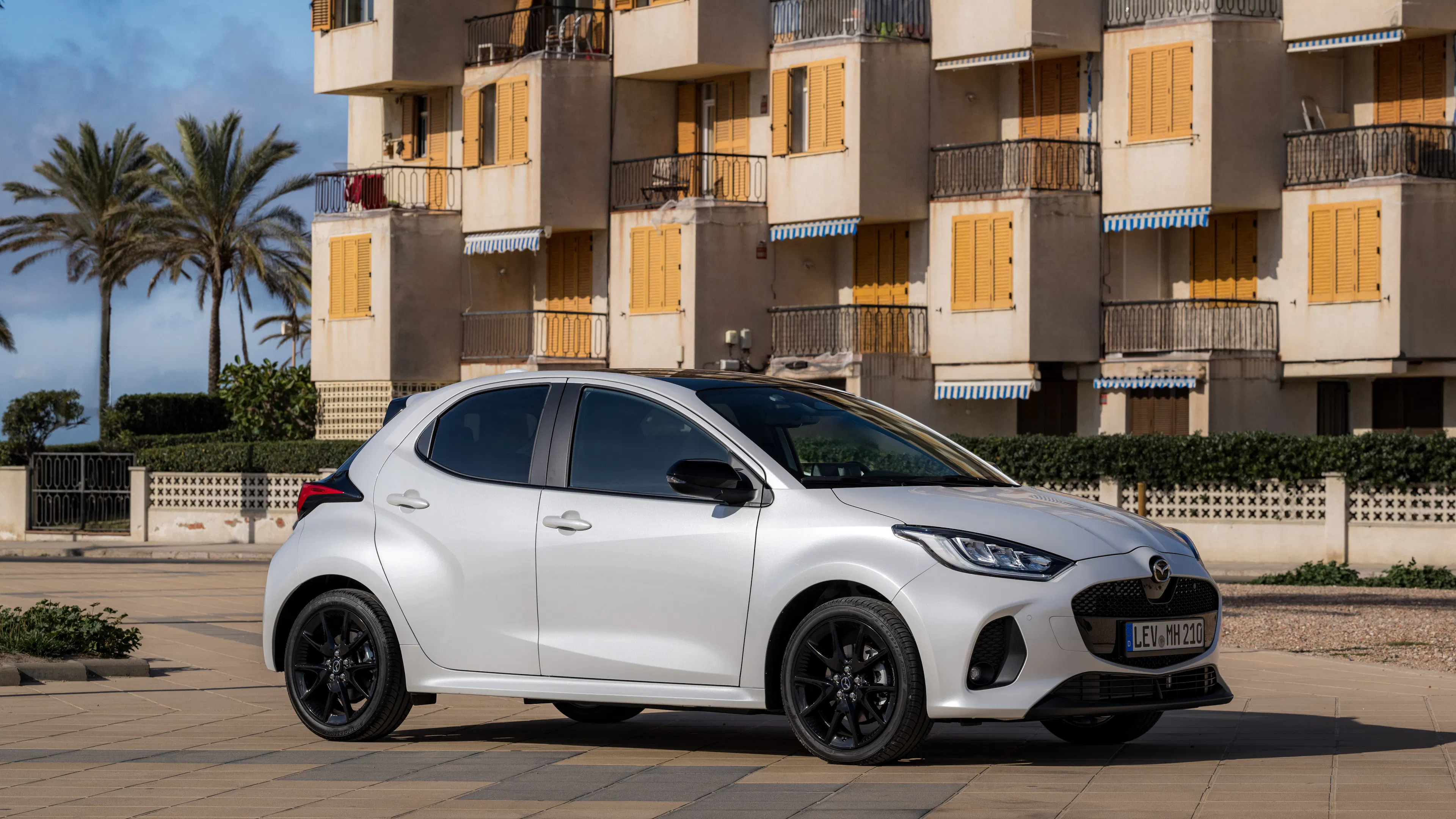 Weißer Mazda2 Hybrid mit schwarzem Dach und schwarzen Leichtmetallfelgen vor einem Wohnhaus mit gelben Fensterläden und Palmen.