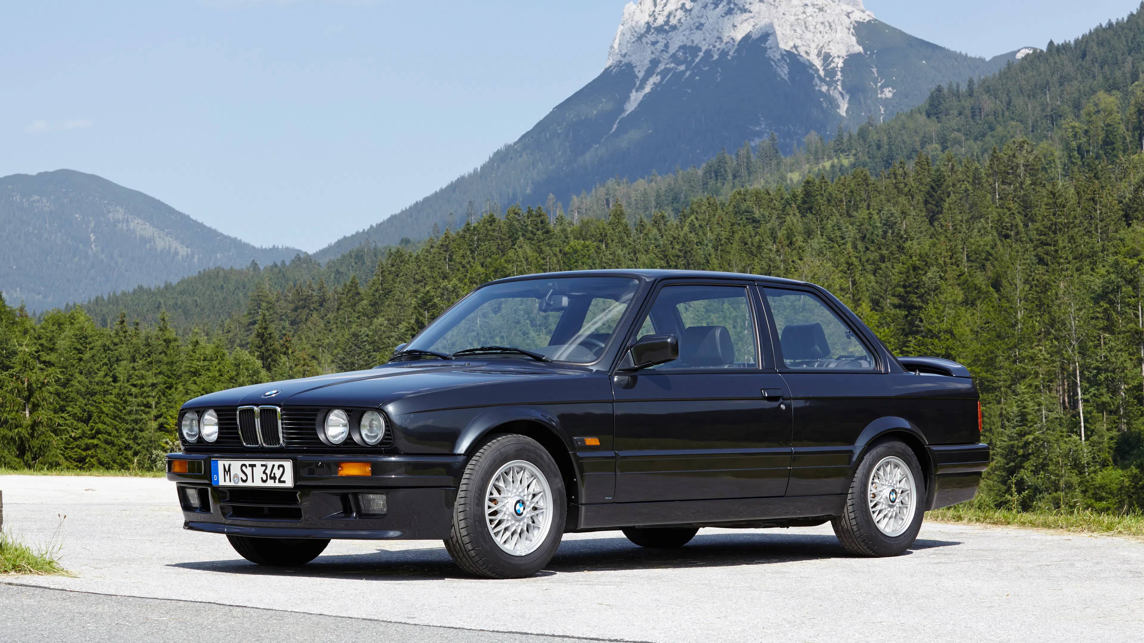 Ein schwarzer BMW 3er E30 steht vor einem Bergpanorama.
