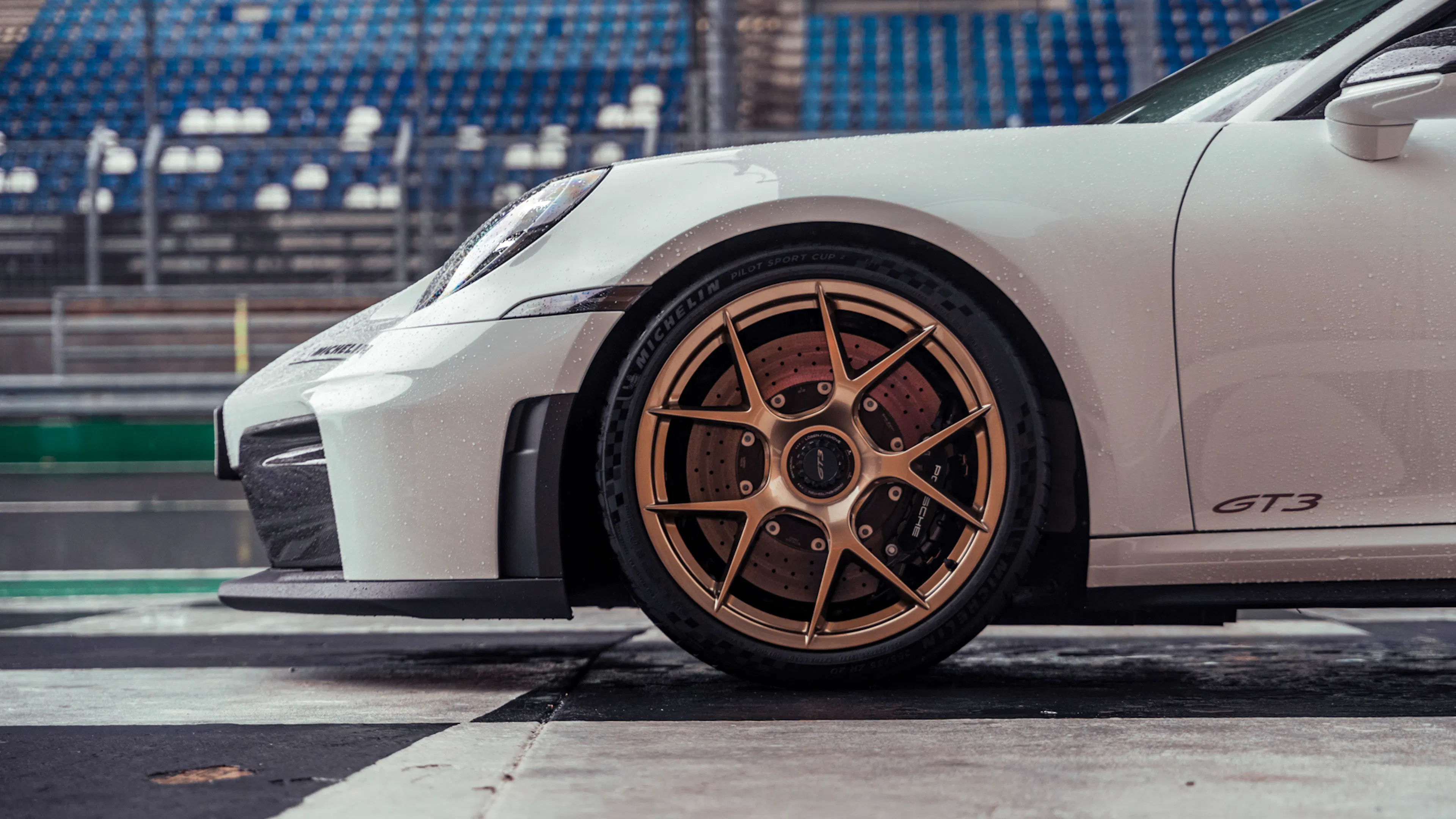 Nahaufnahme des vorderen Fahrzeugbereichs eines Porsche 911 GT3 mit goldfarbenem Leichtmetallrad, gelochter Bremsscheibe, Bremssattel und GT3-Schriftzug bei Regen.
