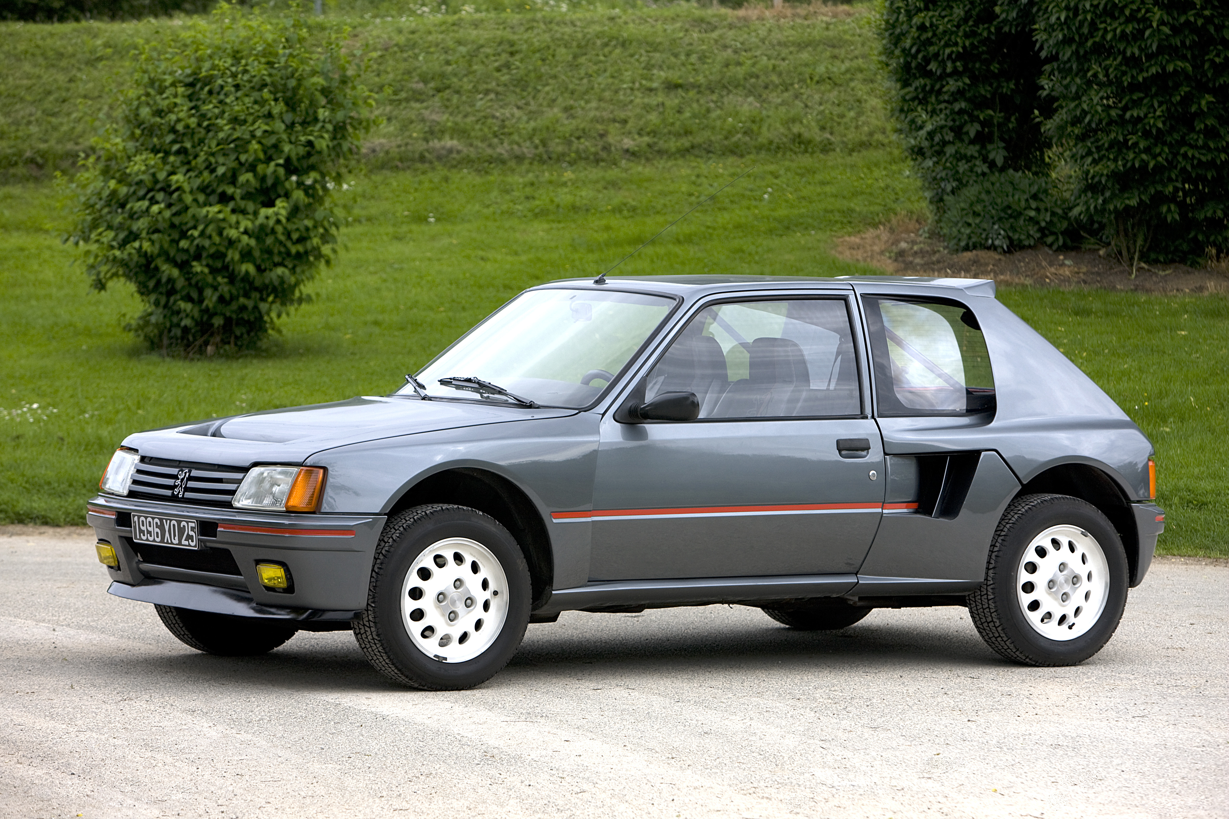 Grauer Peugeot 205 Turbo 16 mit roter Seitenlinie, hellen Alufelgen und Lufteinlass vor dem Hinterrad auf einem Kiesplatz.