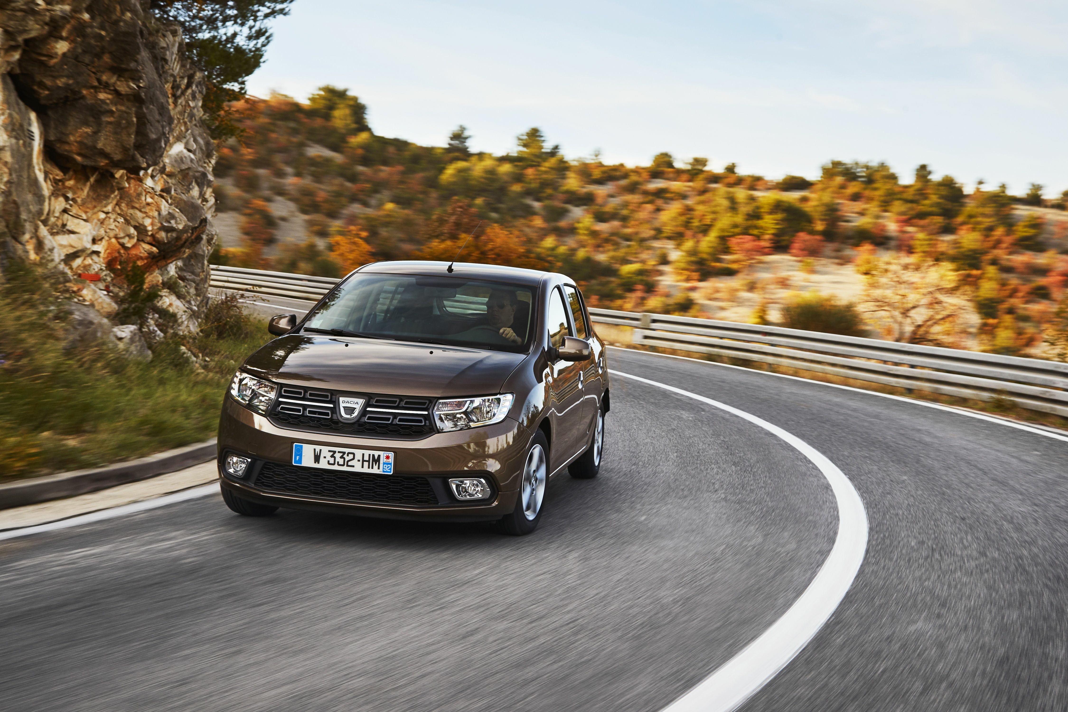 Brauner Dacia Sandero fährt durch eine Kurve einer Landstraße, im Hintergrund eine Felswand und herbstlich gefärbte Büsche.