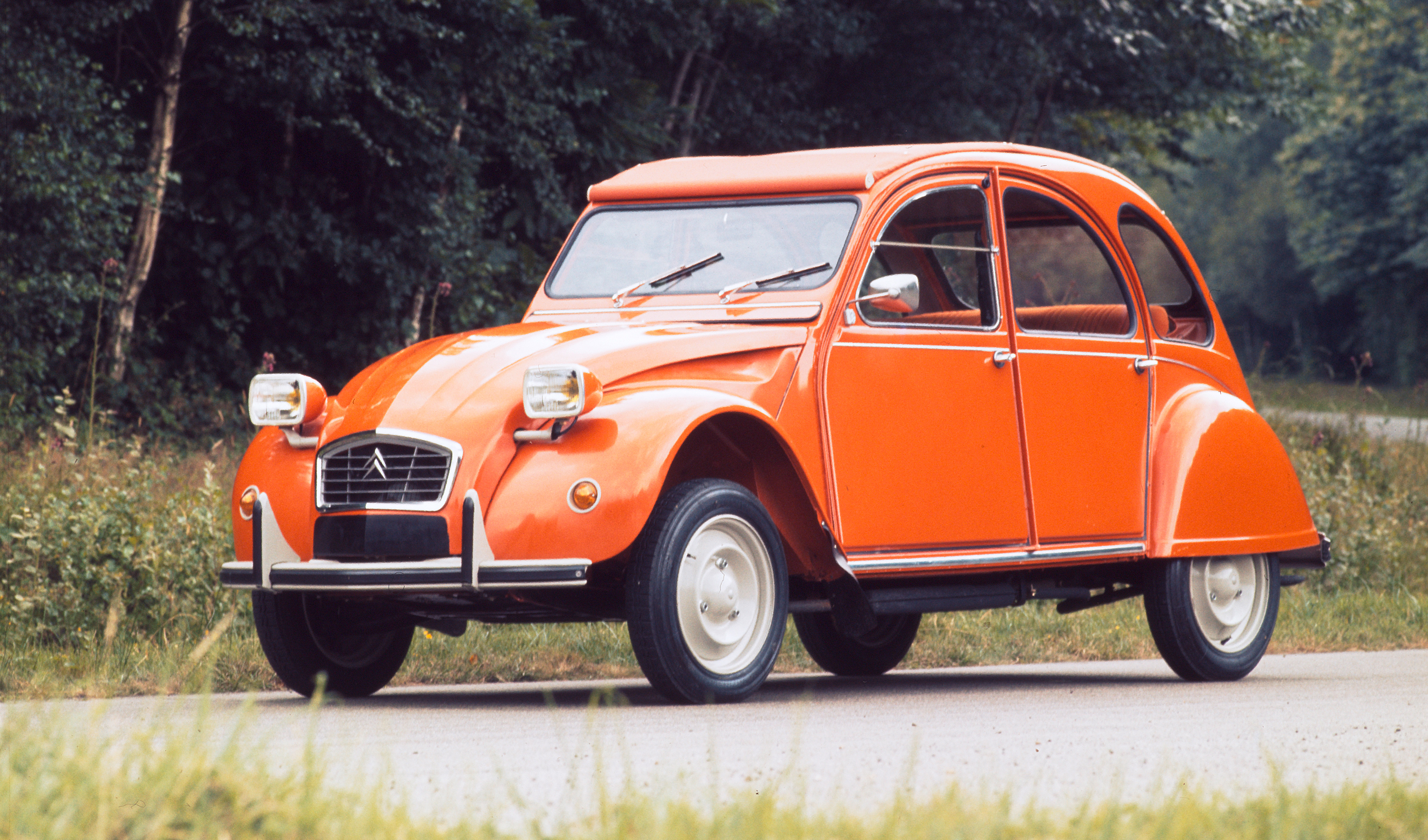Orangefarbener Citroën 2CV6 steht seitlich-frontal auf einer Landstraße vor grüner Hecke, statische Aufnahme