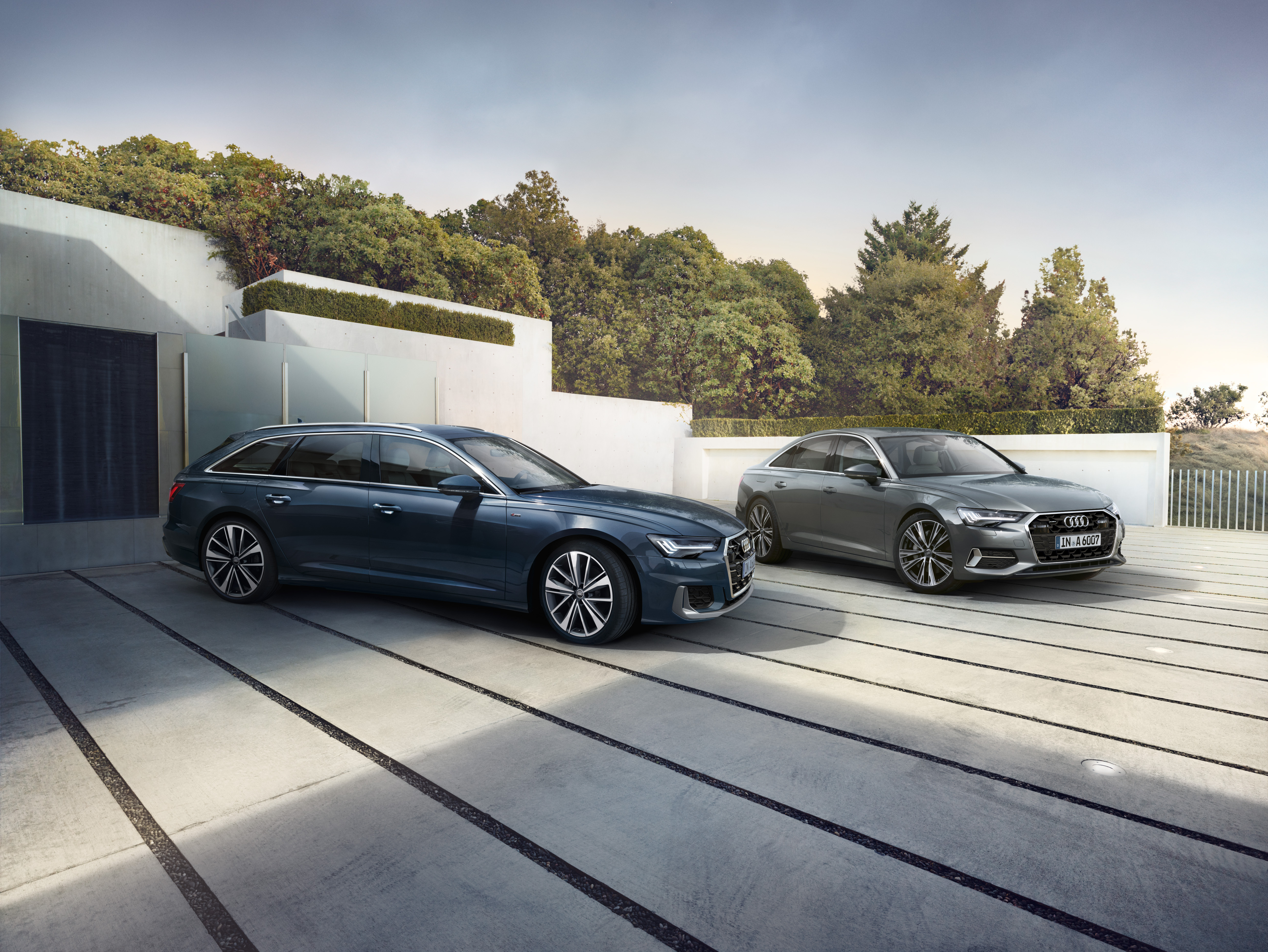 Graue Audi-A6-C8-Limousine und blauer A6 Avant C8 nebeneinander parkend vor einer Gartenmauer mit Hecke.