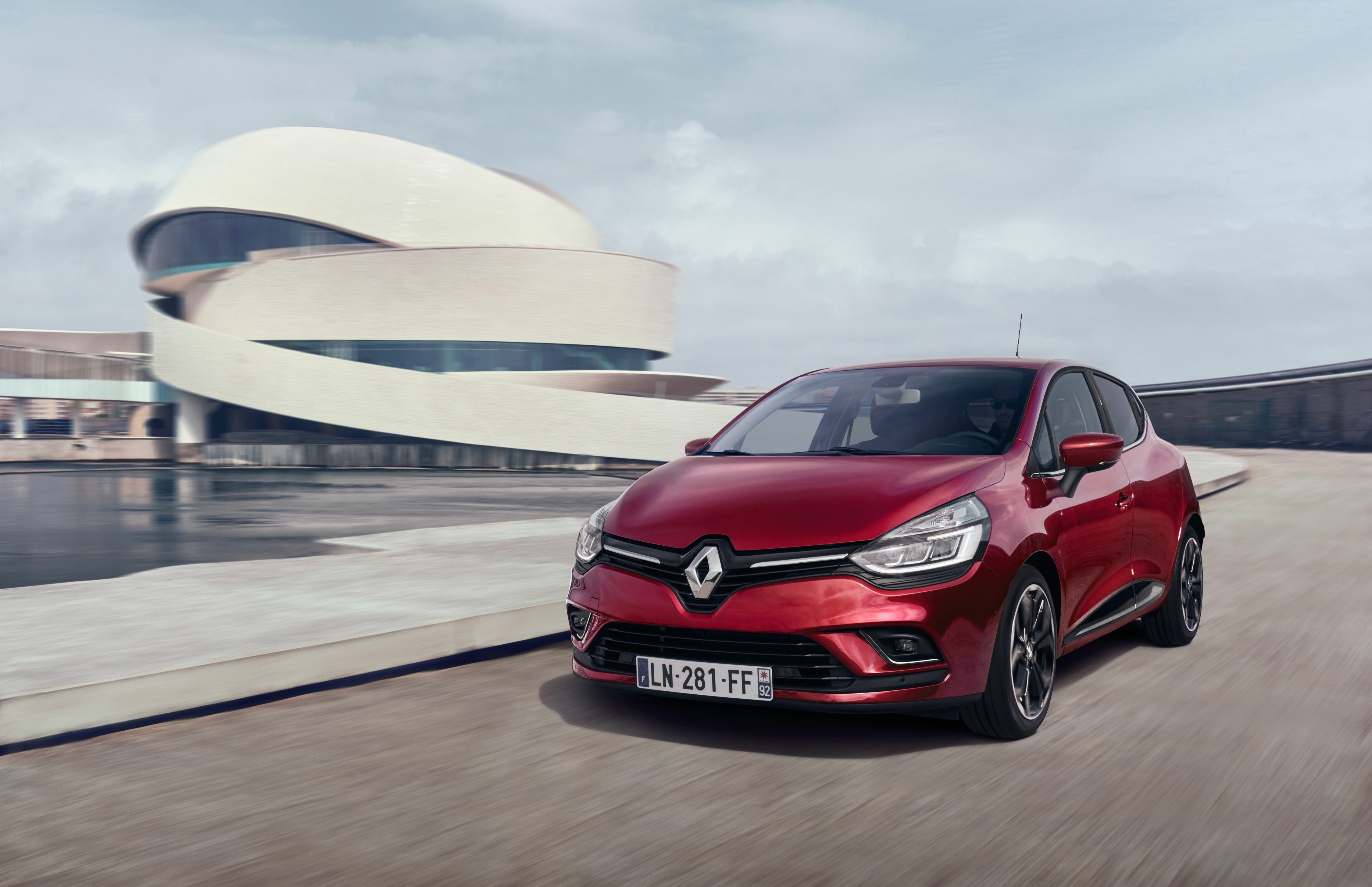 Roter Renault Clio fährt vor einem weißen, geschwungenen Gebäude am Wasser.