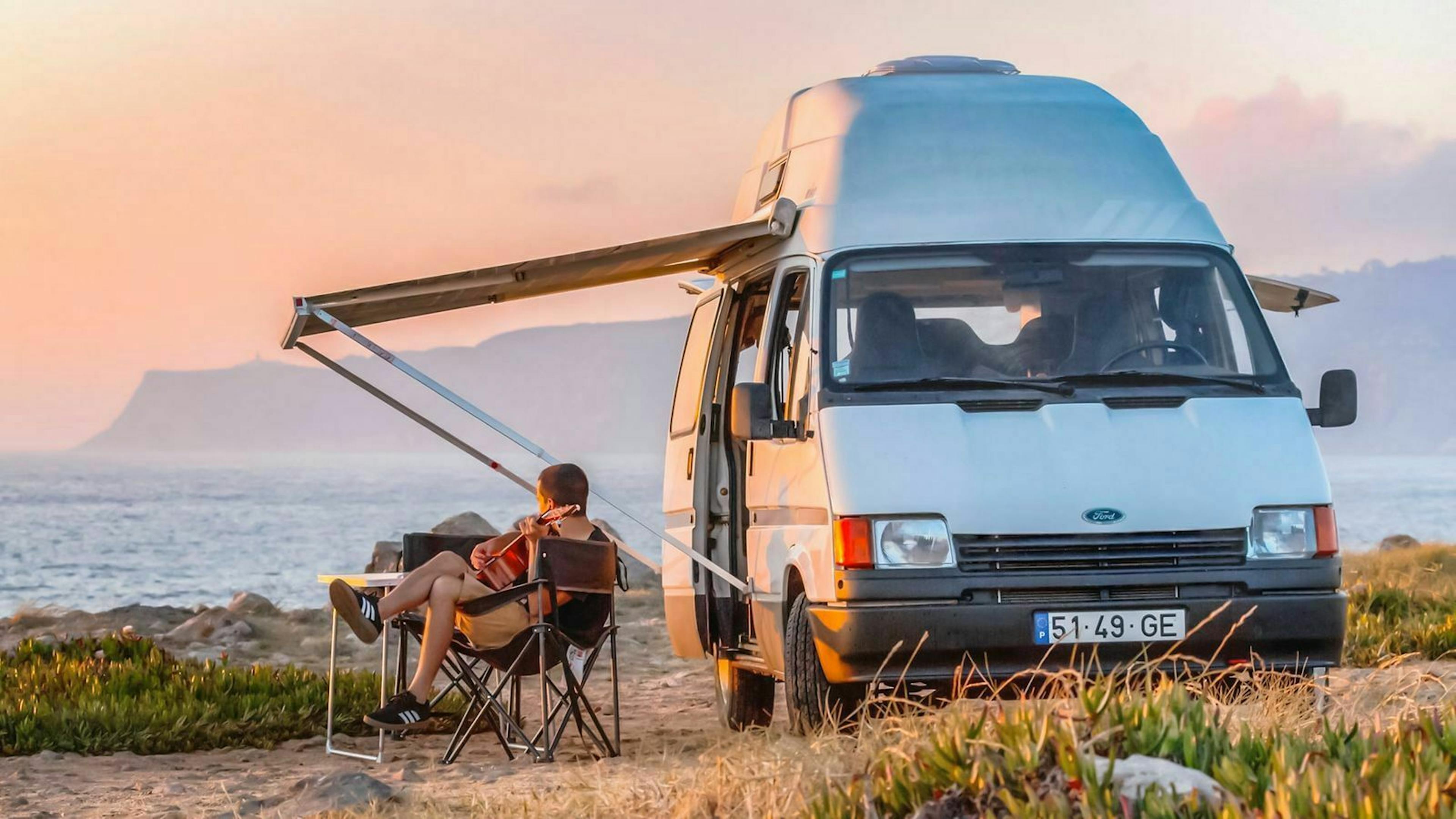 Ein junger Mann beobachtet an einer Küste den Sonnenuntergang und sitzt vor seinem Wohnmobil.