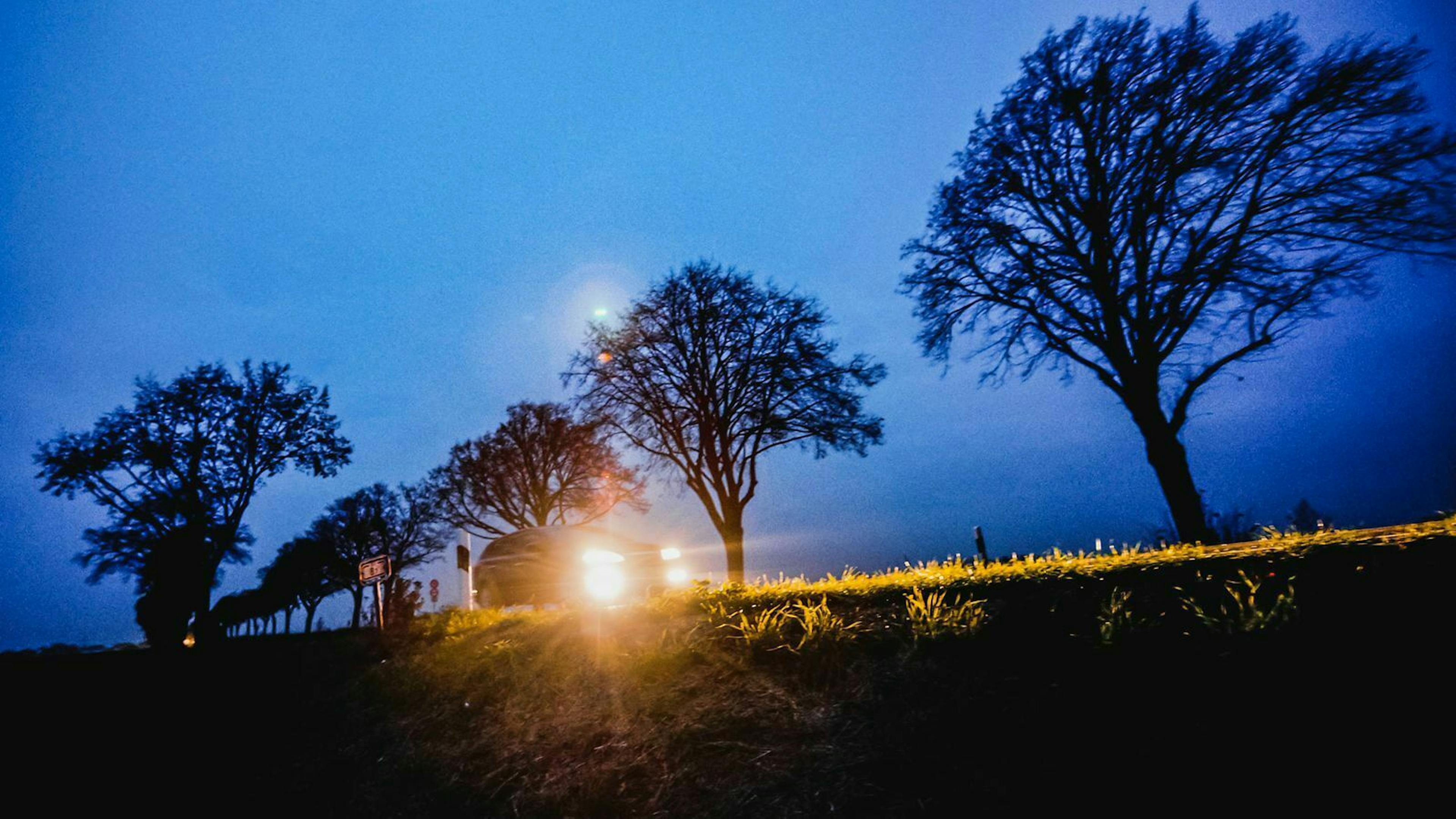 Ein Auto fährt in der Dämmerung mit eingeschalteten Scheinwerfern über eine Landstraße.