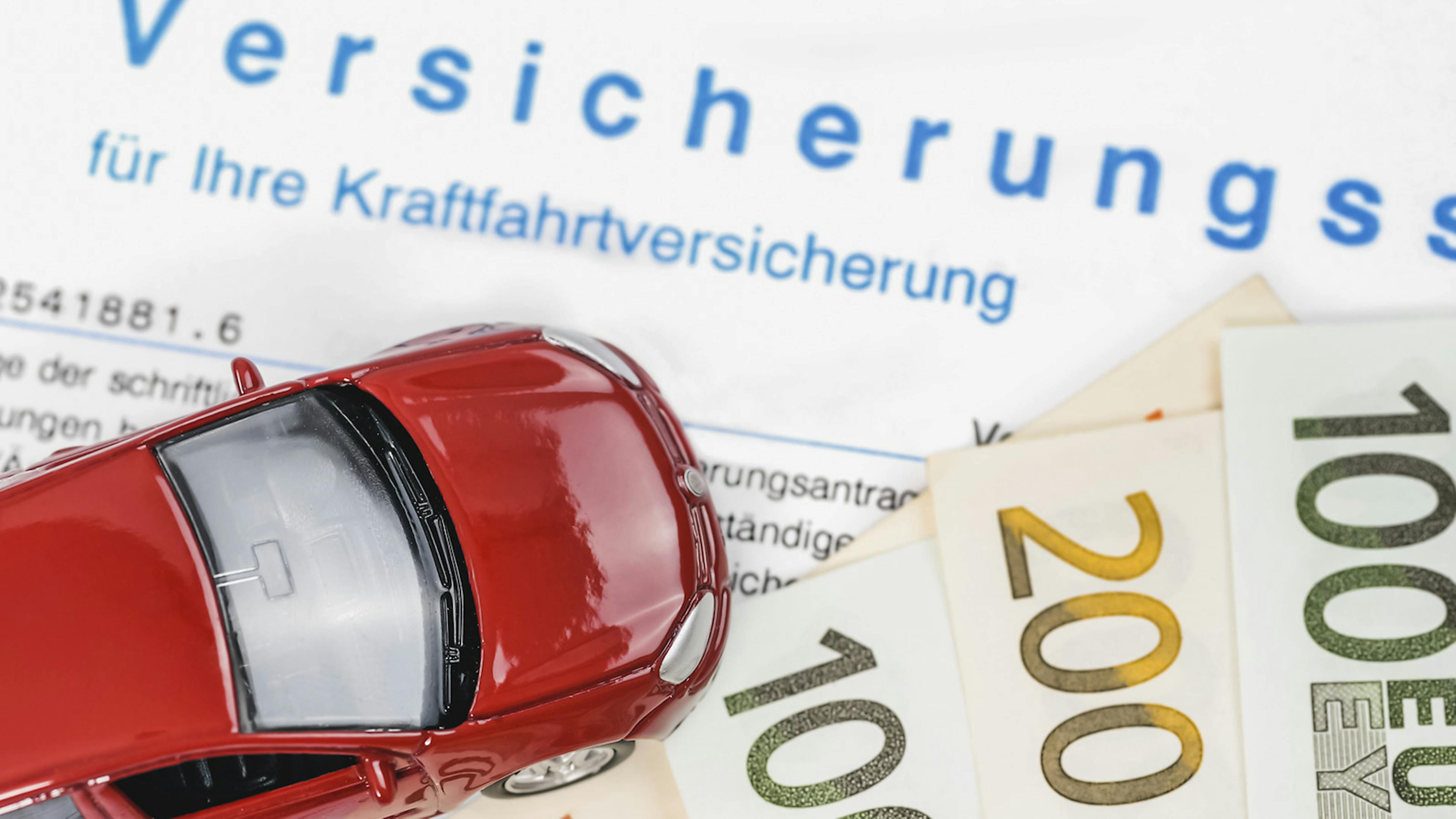 Ein rotes Modellauto steht auf einem Geldschein und einem Blatt Papier, auf dem in blauer Schrift "Versicherungsschein" steht.
