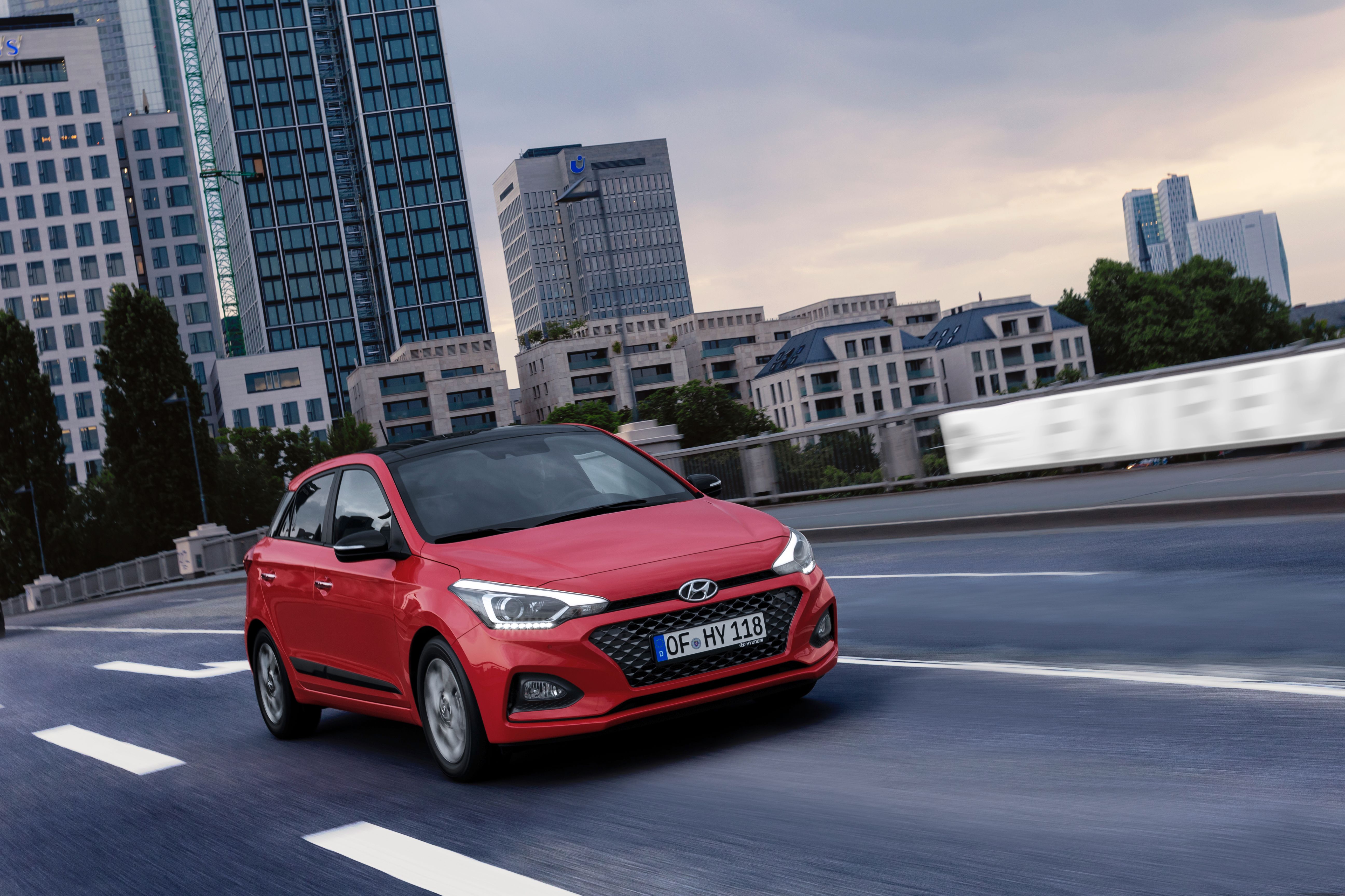 Roter Hyundai i20 mit schwarzem Dach fährt in der Dämmerung vor Hochhäusern über eine Stadtstraße.