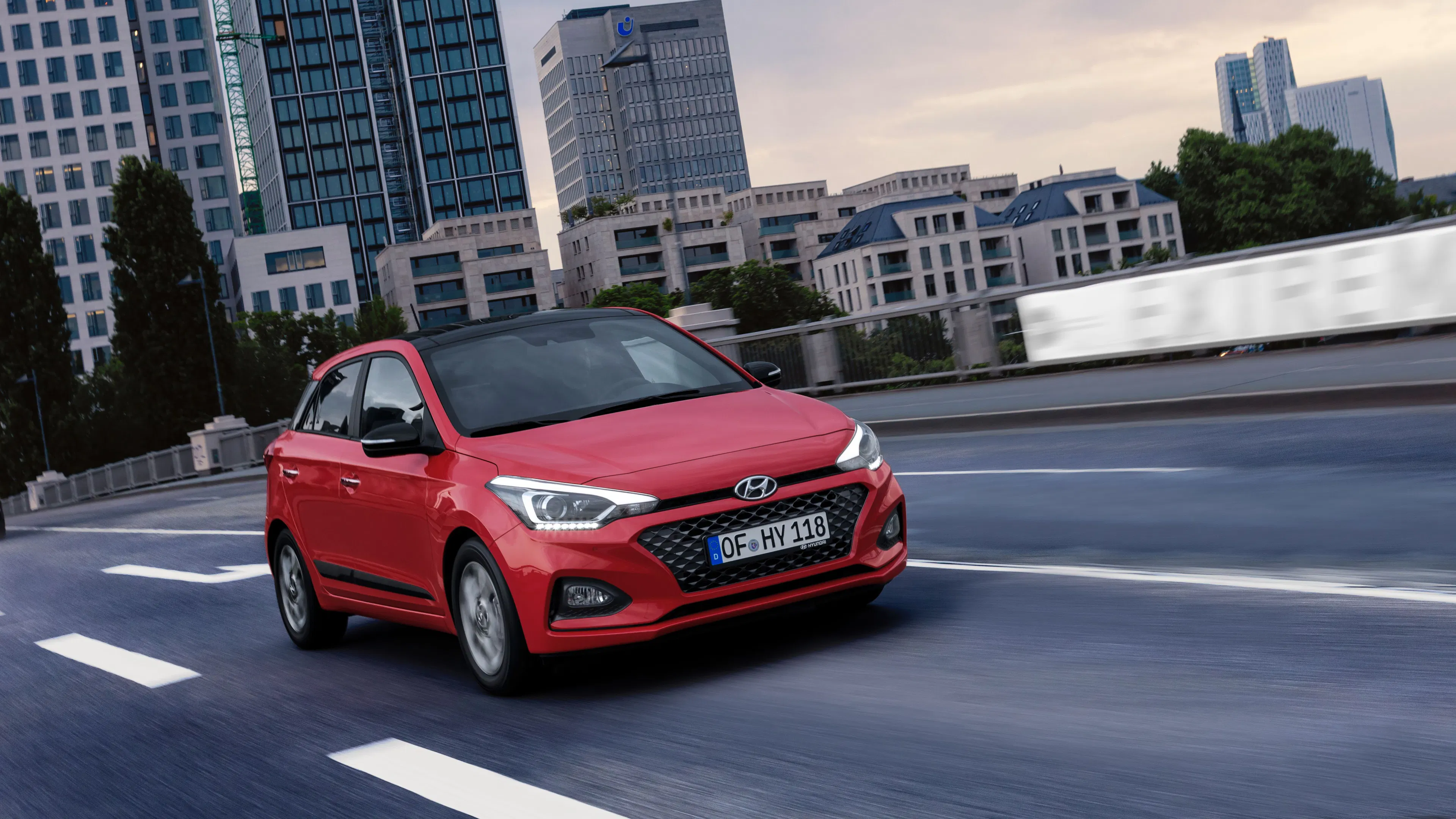 Roter Hyundai i20 mit schwarzem Dach fährt in der Dämmerung vor Hochhäusern über eine Stadtstraße.