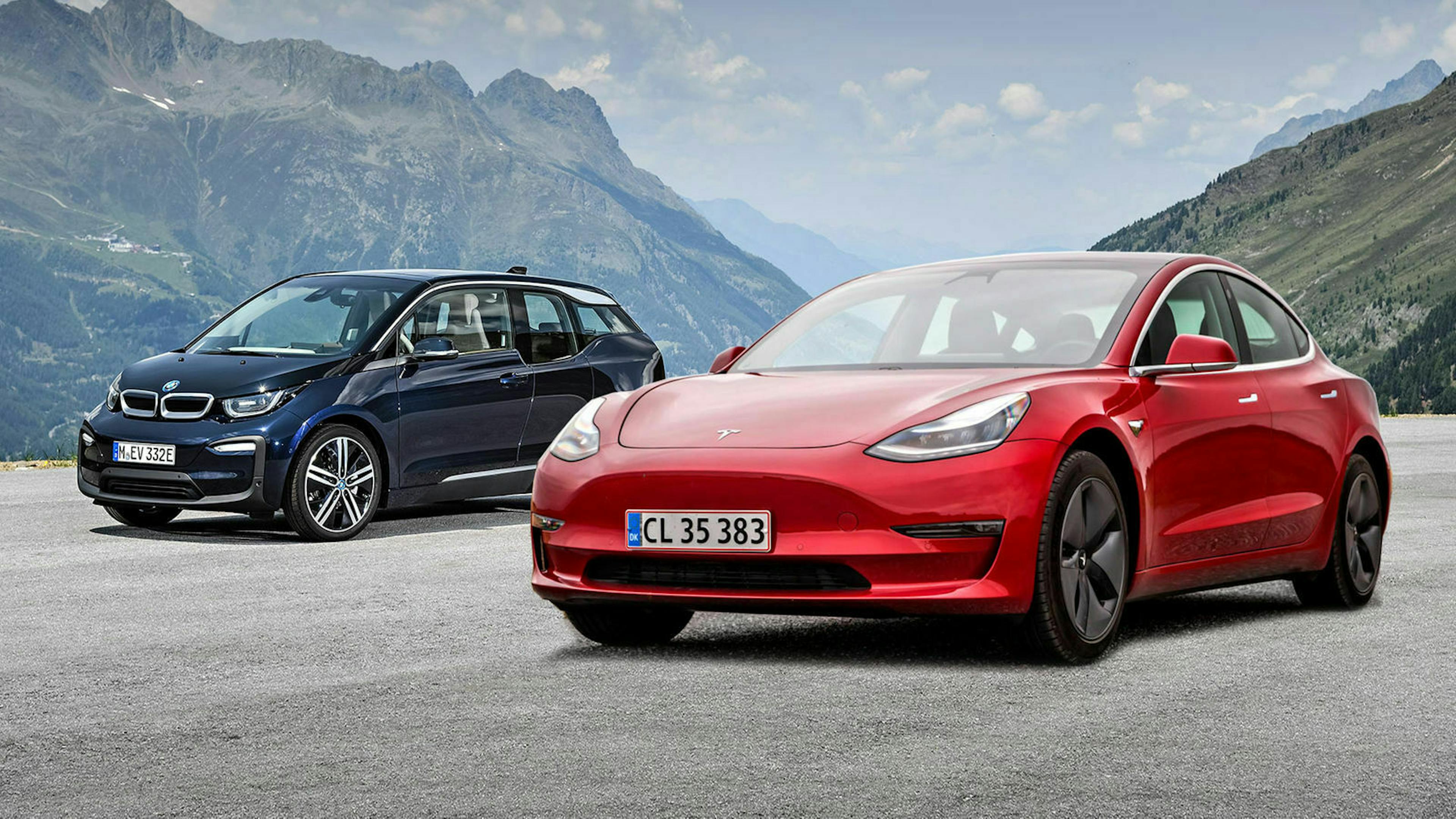 Ein BMW i3 und ein Tesla Model 3 parken auf einer Asphaltfläche vor einem Alpenpanorama.