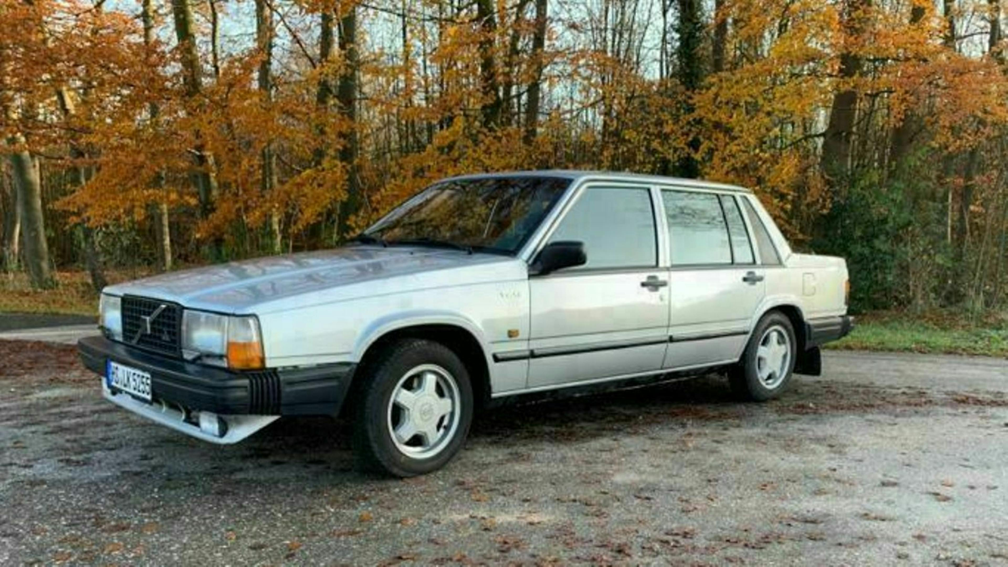 Silberner Volvo 940 Kombi geparkt auf einem Waldweg im Herbst, umgeben von Bäumen mit buntem Laub.