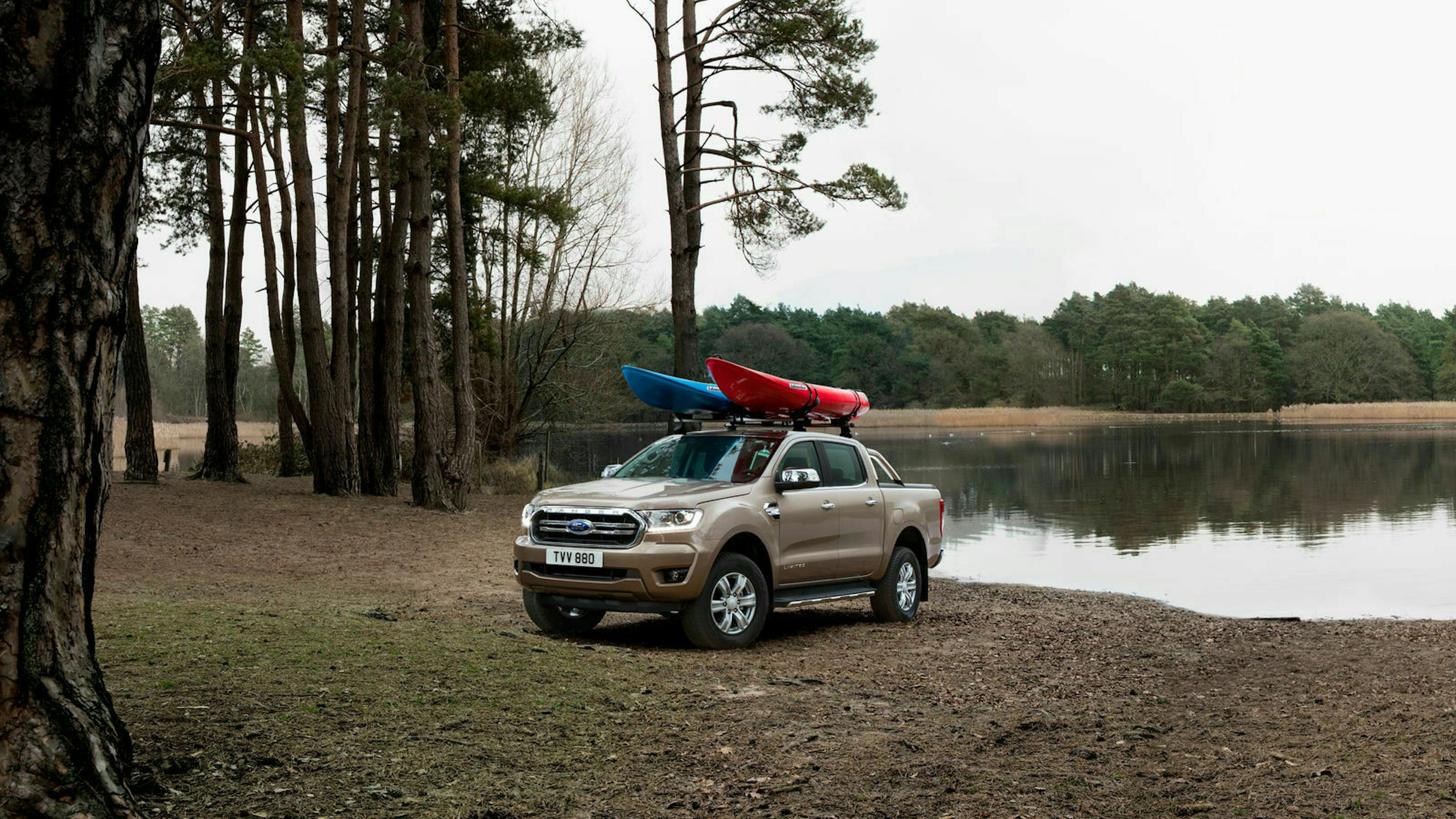 Ein grauer Ford Ranger steht an einem See, auf dem Dach sind ein rotes und ein blaues Kajak befestigt.