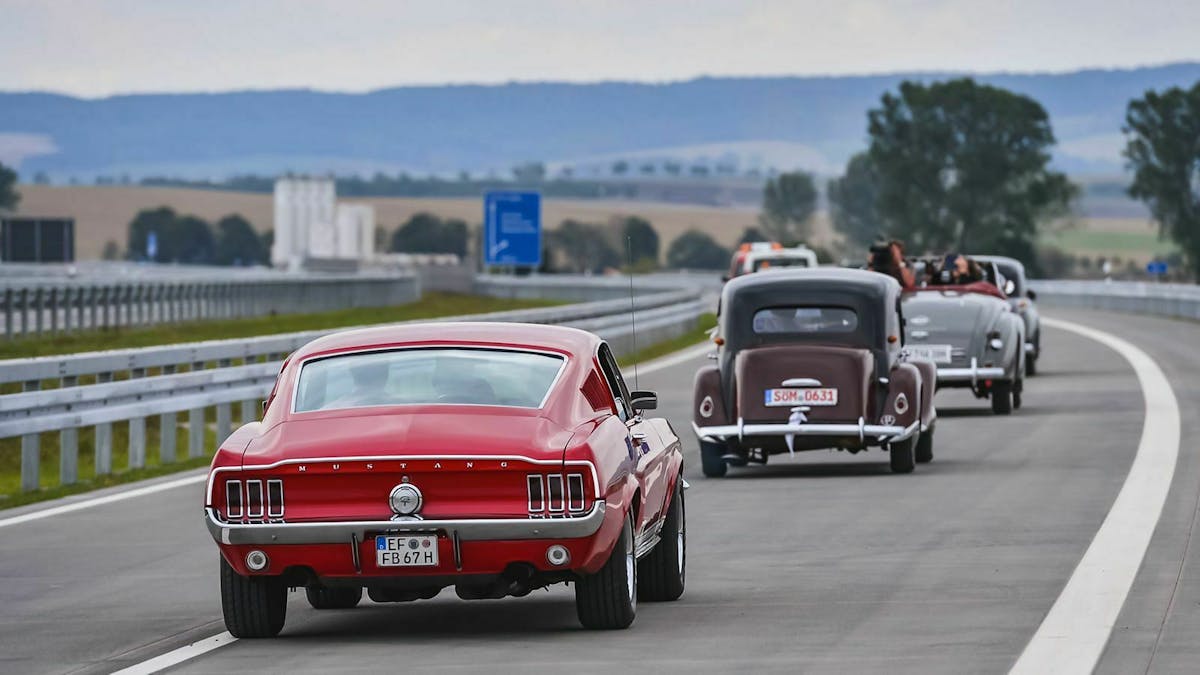 Ein roter Ford Mustang fährt in einer Kolonne älterer Autos über eine Autobahn.