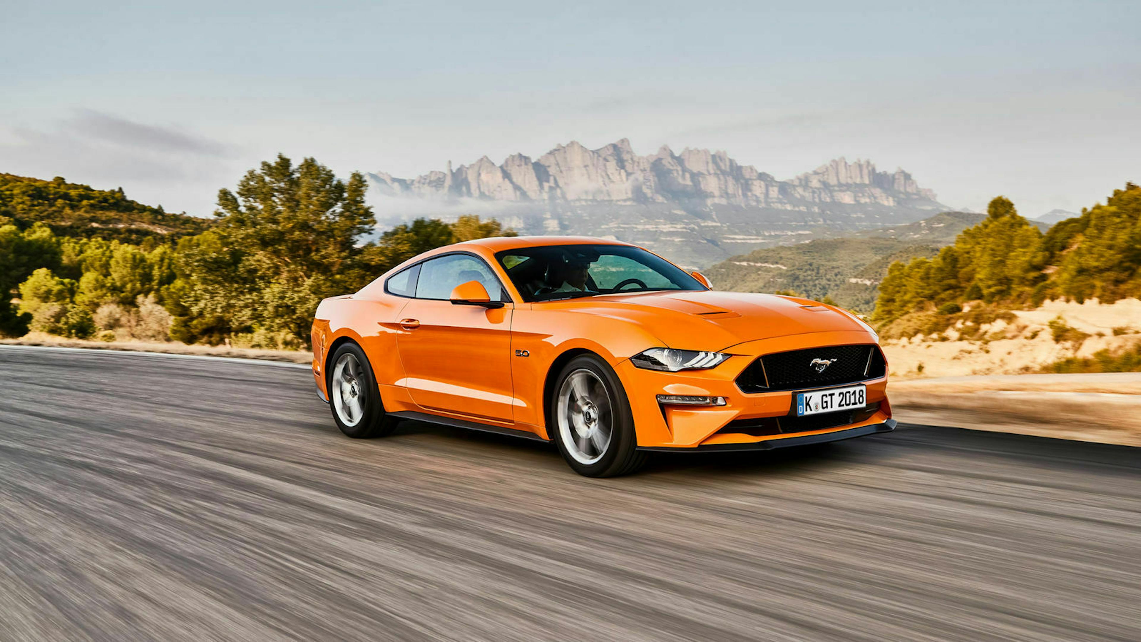 Ein orangefarbener Ford Mustang fährt auf einer Straße durch eine bergige Landschaft