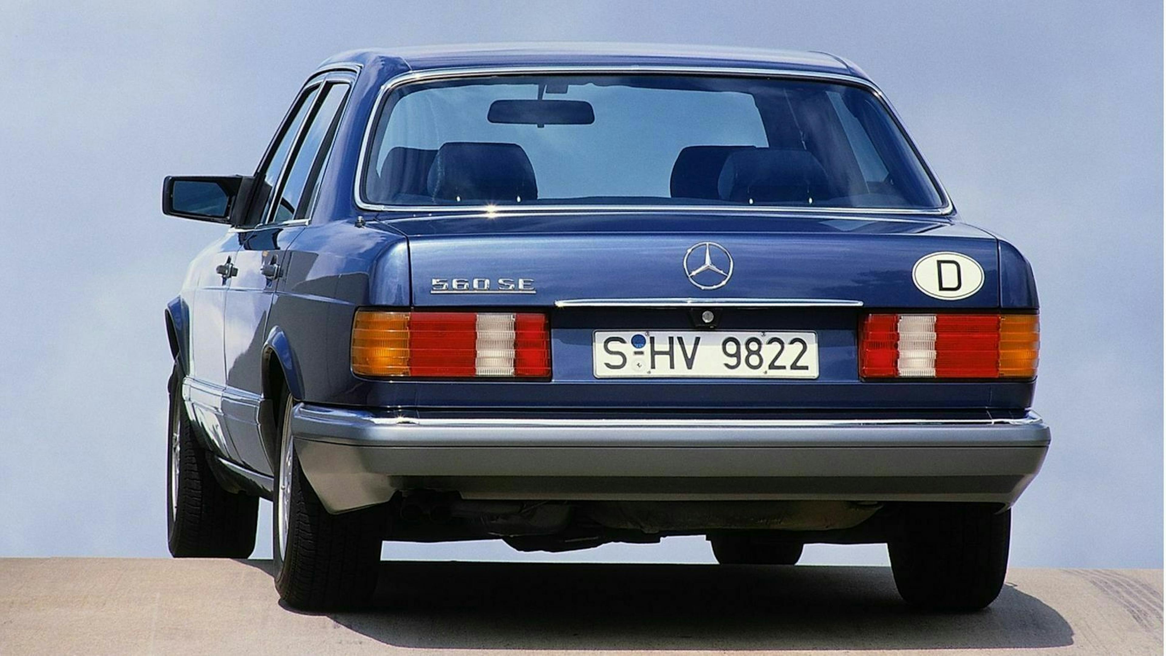 Blauer Mercedes 560 SE von hinten, auf einer Straße mit klarem Himmel. Deutsche Kennzeichen und ovaler Aufkleber "D" sichtbar.