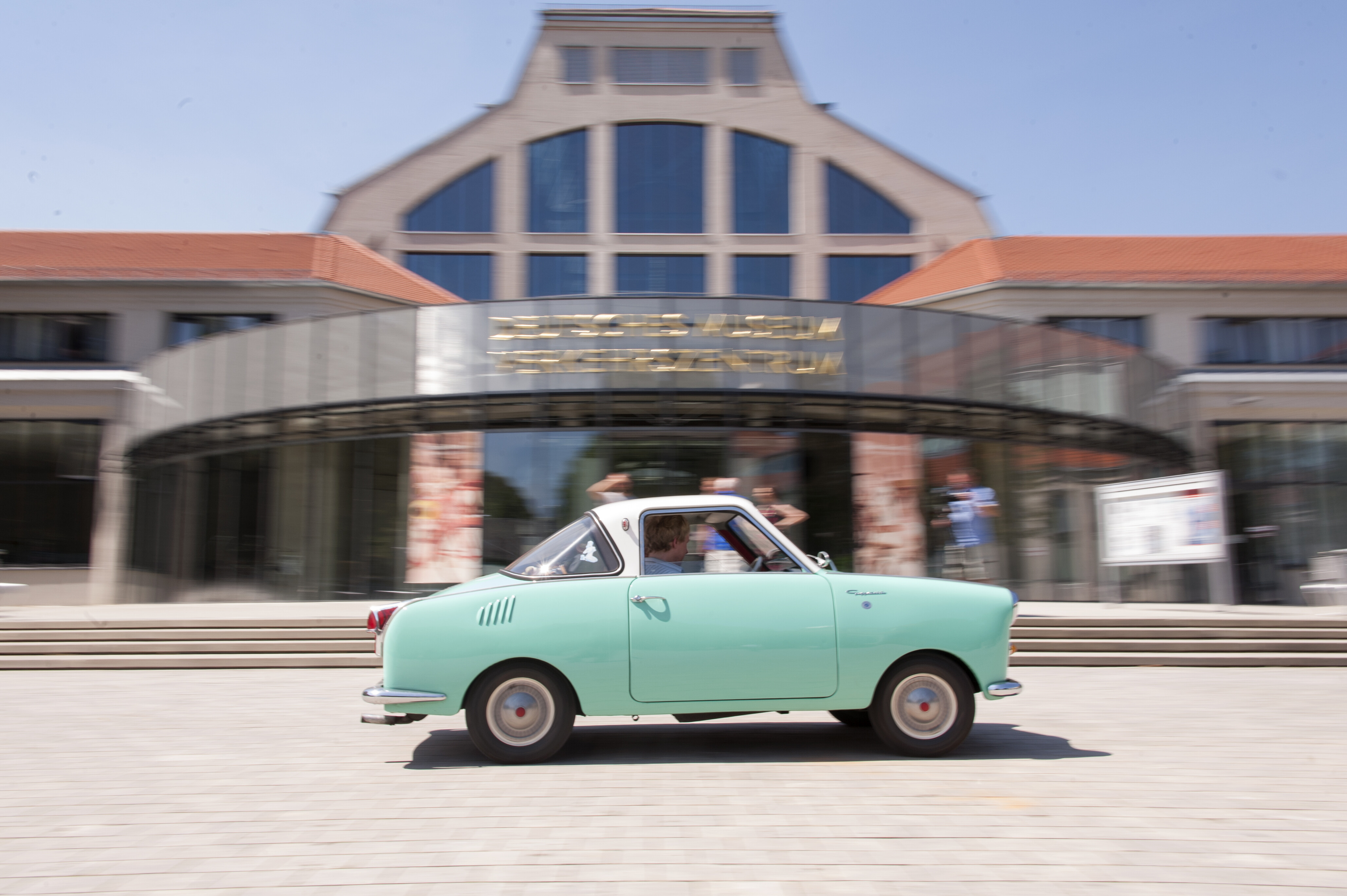 Türkisfarbenes Goggomobil in Fahrt, Seitenansicht vor dem Verkehrszentrum des Deutschen Museums in München.