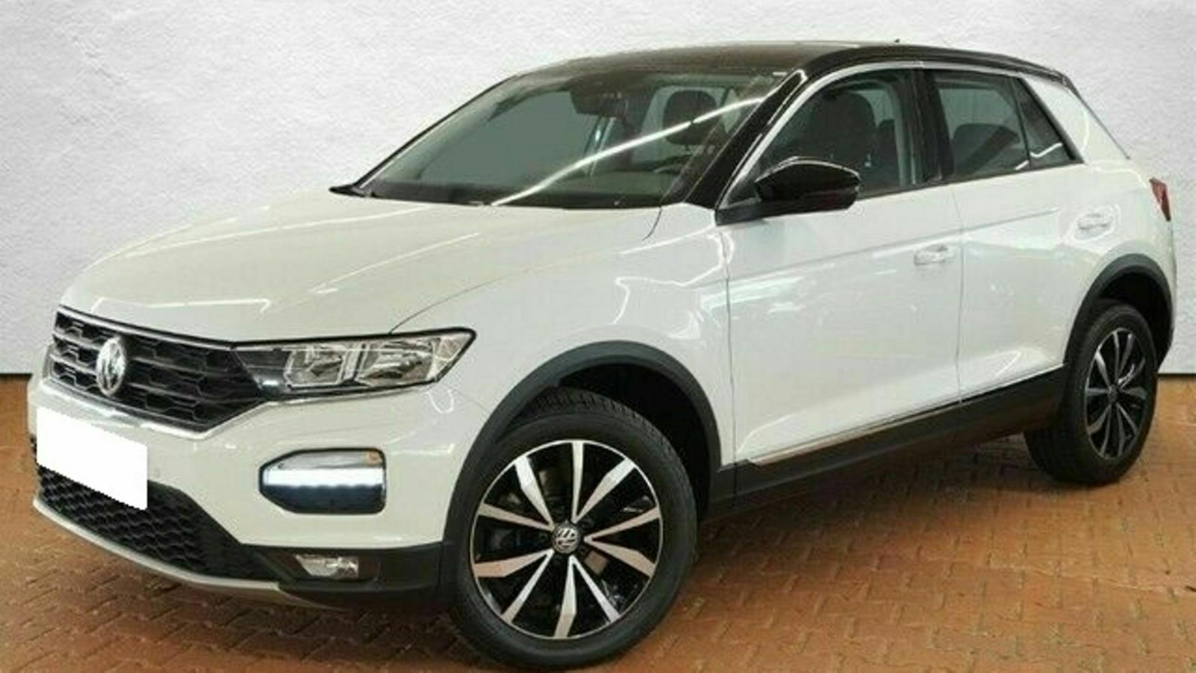 Weißer VW T-Roc SUV mit schwarzen Akzenten und sportlichen Alufelgen, geparkt auf gepflastertem Boden vor einer weißen Wand.