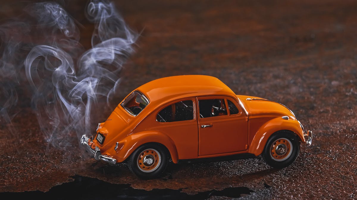 Ein Modellauto vom typ VW Käfer steht auf rostigem Blech. Es qualmt unter dem Auto hervor.
