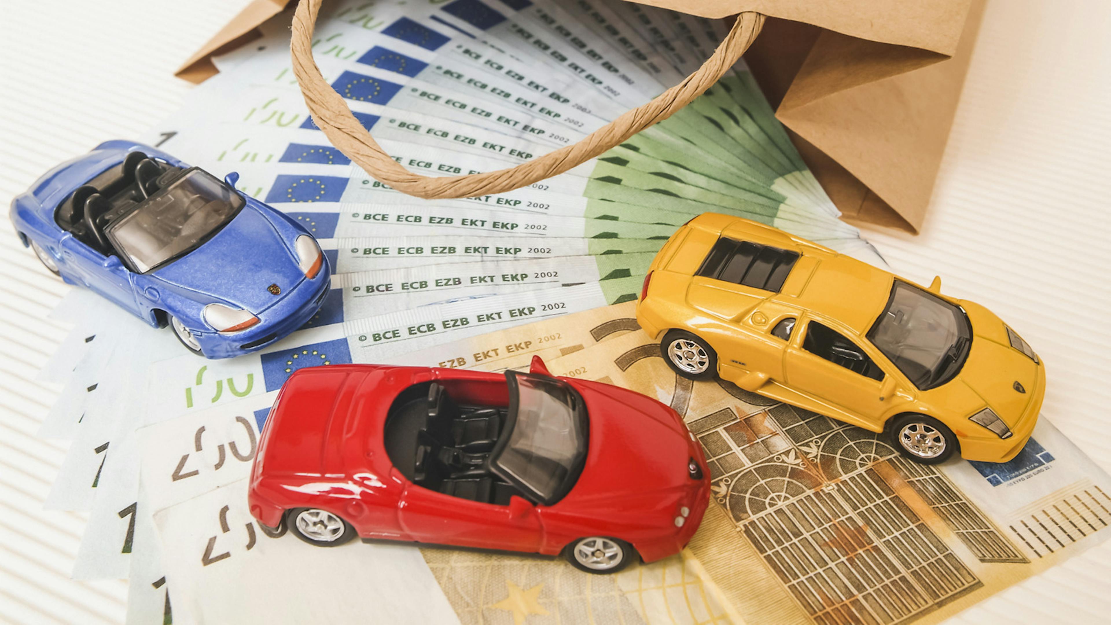 Aus einer braunen Papiertüte läuft ein Fächer aus 100- und 50-Euro-Banknoten, auf denen drei Spielzeugautos platziert sind.