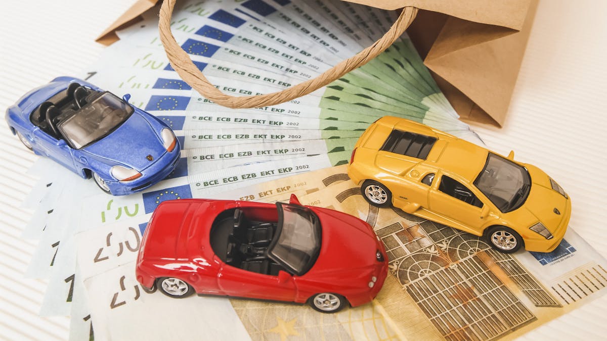 Aus einer braunen Papiertüte läuft ein Fächer aus 100- und 50-Euro-Banknoten, auf denen drei Spielzeugautos platziert sind.
