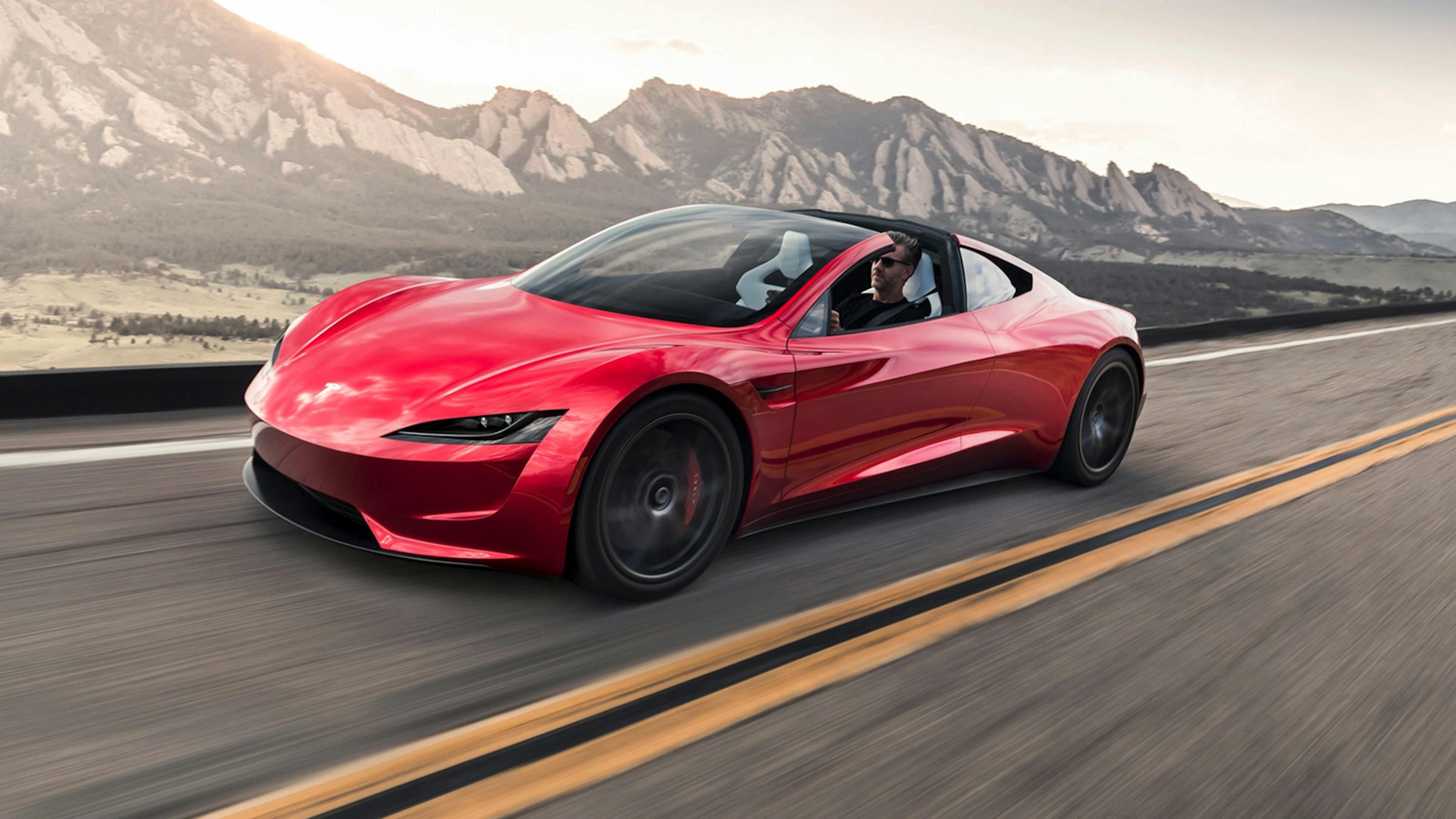 Ein roter Tesla Roadster fährt vor einem Bergpanorama über eine Landstraße