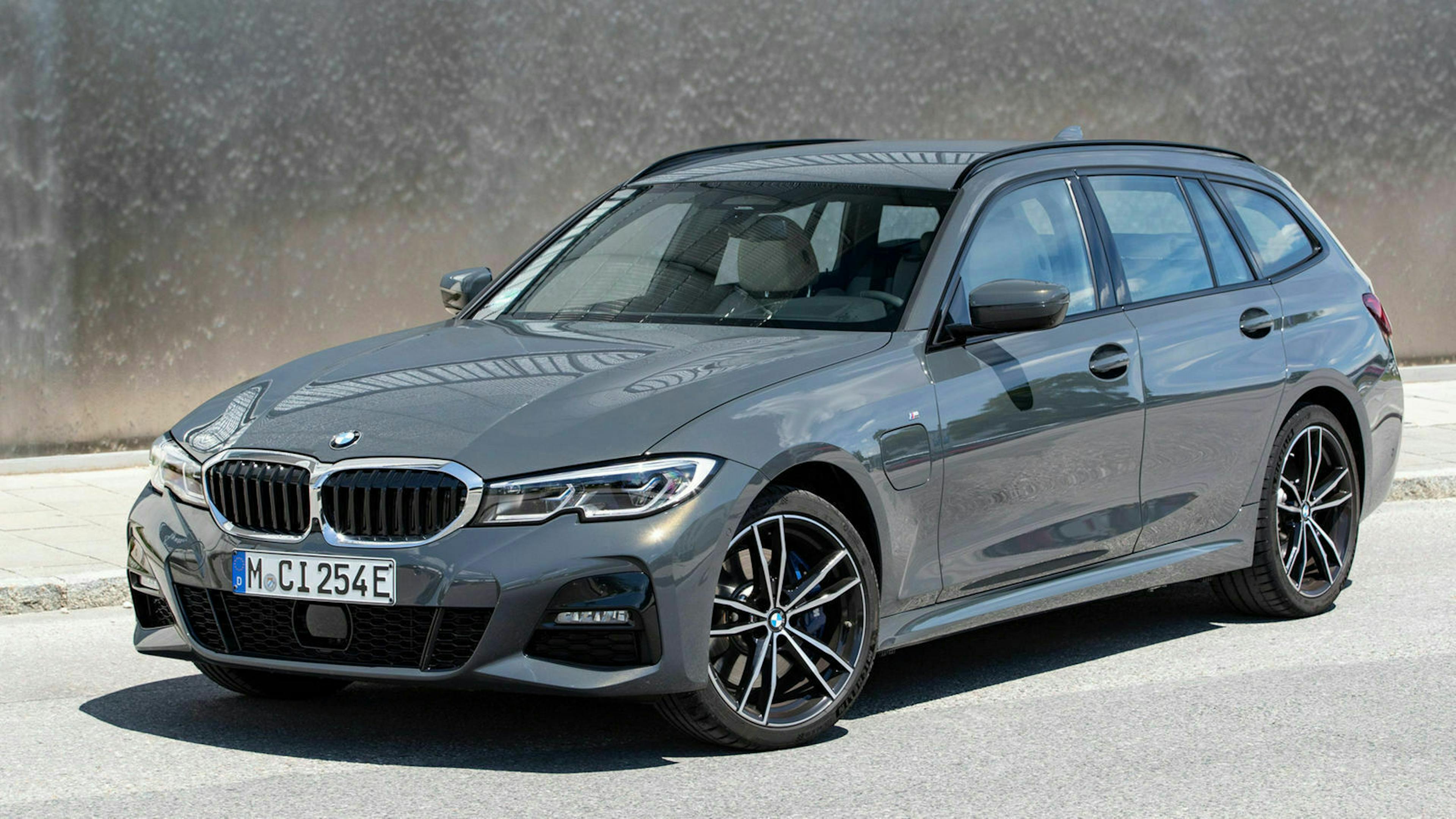 Ein grauer BMW 320e Touring steht vor einer dunklen Betonwand. 