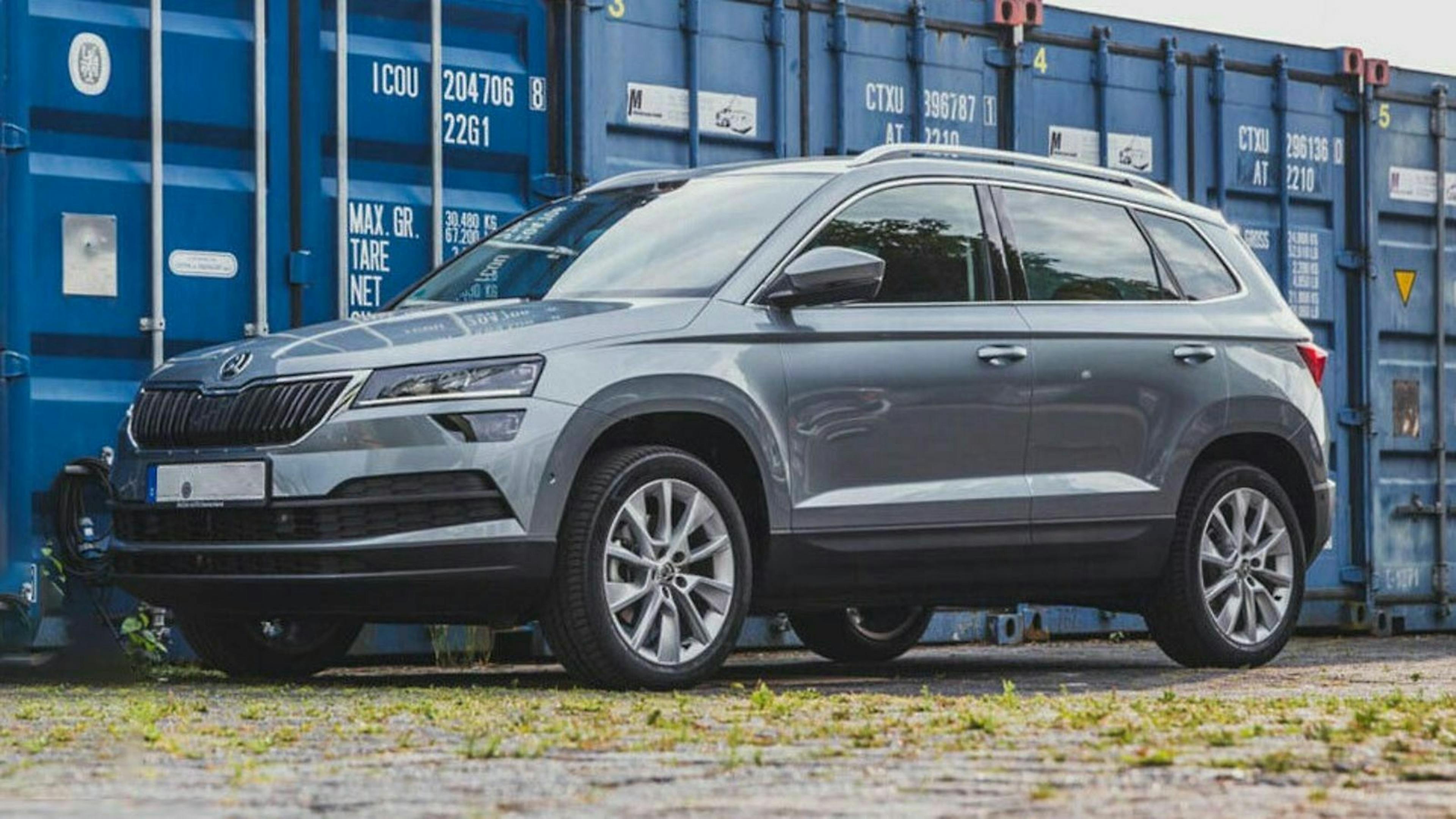 Silberner SUV geparkt vor blauen Containern auf einem gepflasterten Untergrund, seitliche Ansicht.
