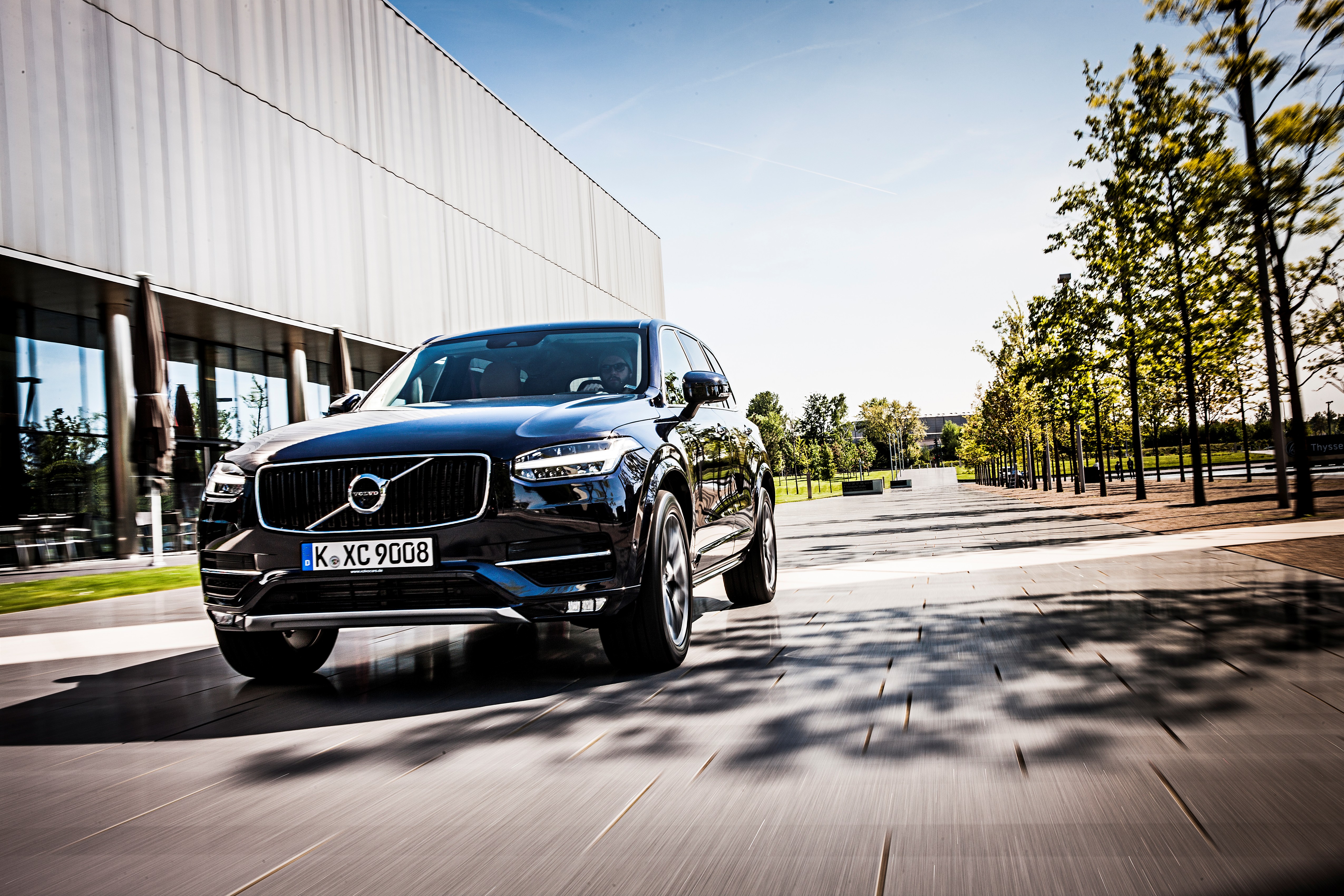 Ein schwarzer Volvo XC90 fährt vor einem modernen Gebäude über eine Straße.