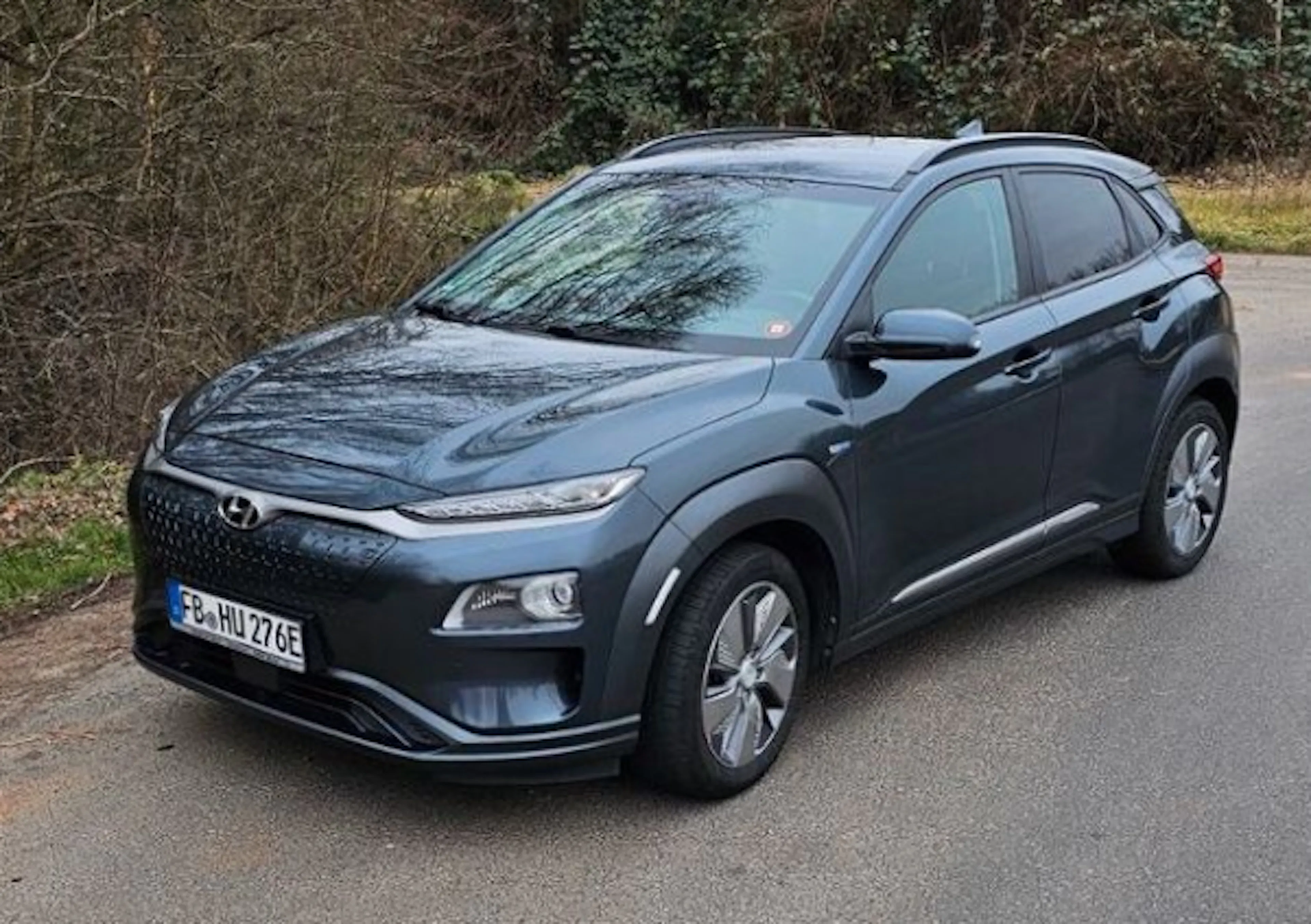 Ein grauer Hyundai Kona Electric steht auf einer Straße, umgeben von Bäumen. Das Auto hat ein modernes Design und glänzende Räder.