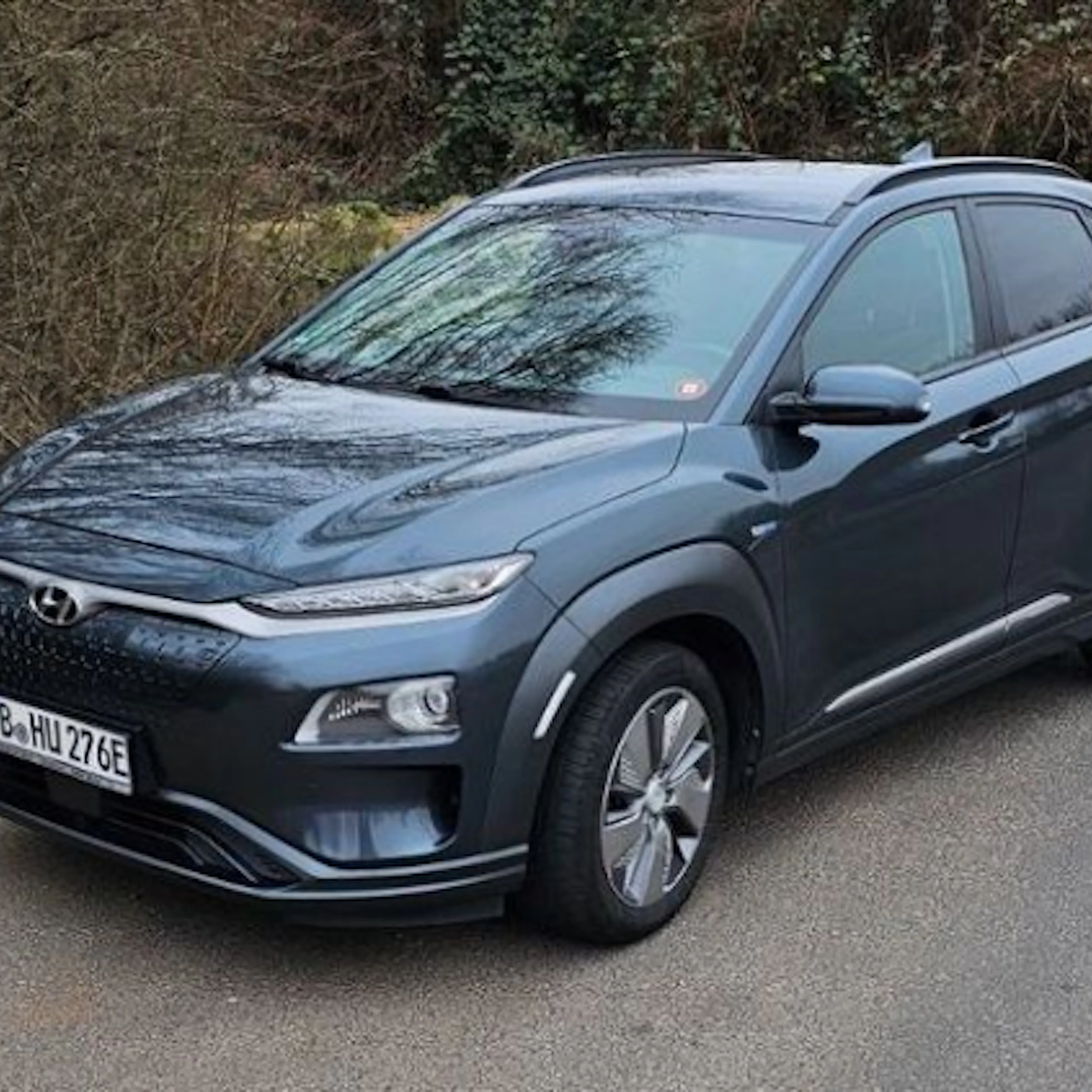 Ein grauer Hyundai Kona Electric steht auf einer Straße, umgeben von Bäumen. Das Auto hat ein modernes Design und glänzende Räder.