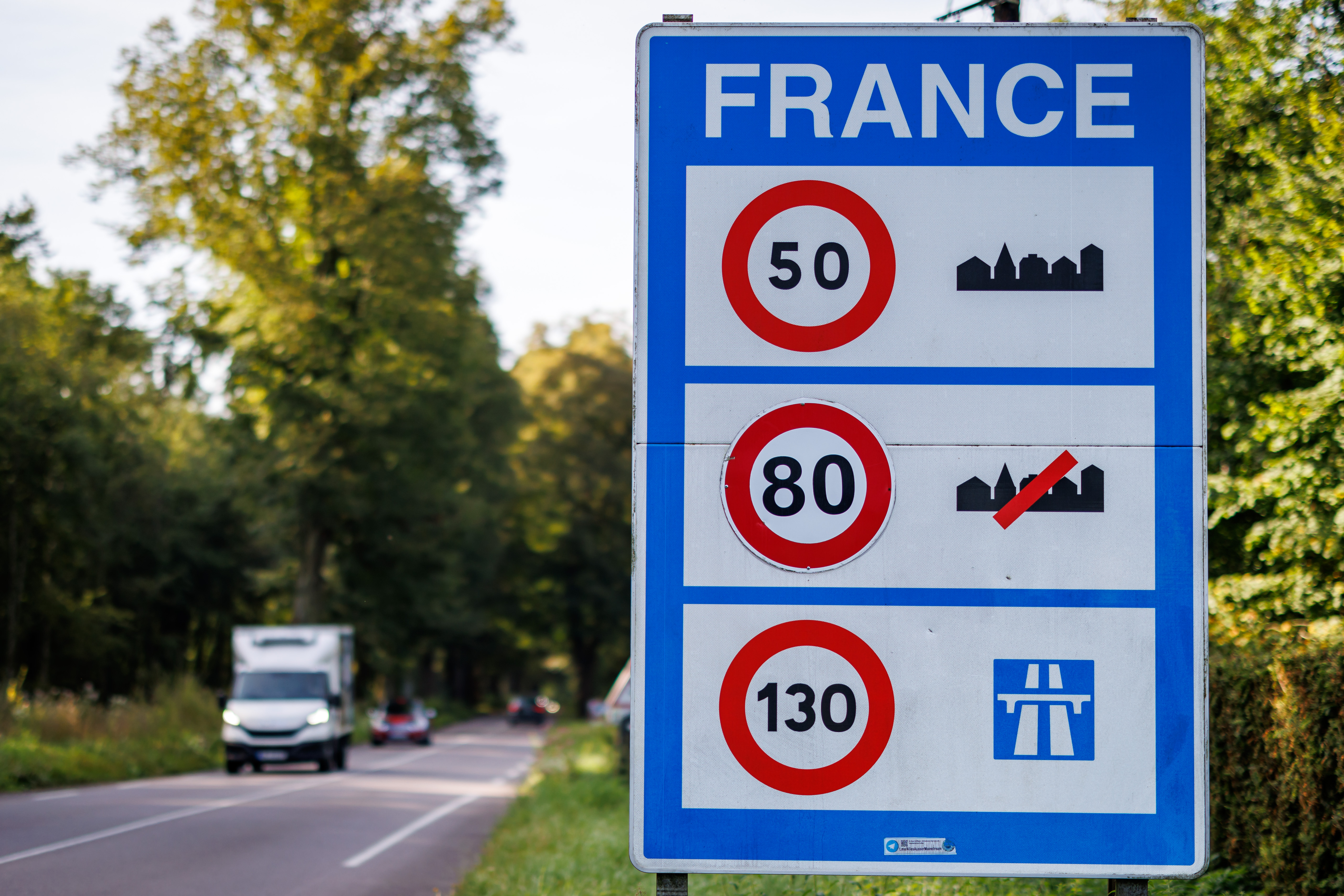 A blue traffic sign on a road informs about the speed limits in France. It shows that a speed limit of 50 km/h applies in built-up areas, 80 km/h outside built-up areas and 130 km/h on motorways.