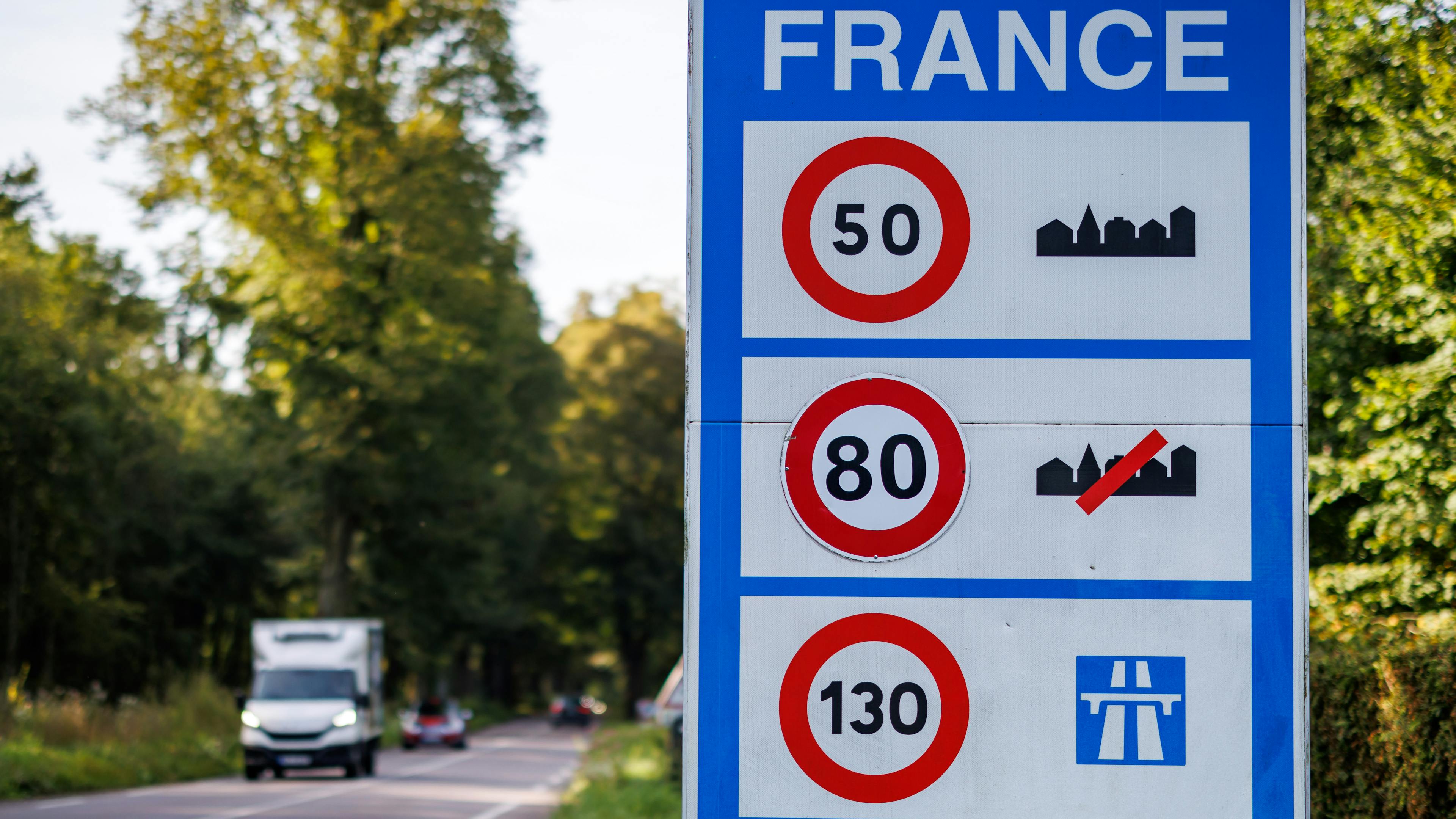 A blue traffic sign on a road informs about the speed limits in France. It shows that a speed limit of 50 km/h applies in built-up areas, 80 km/h outside built-up areas and 130 km/h on motorways.