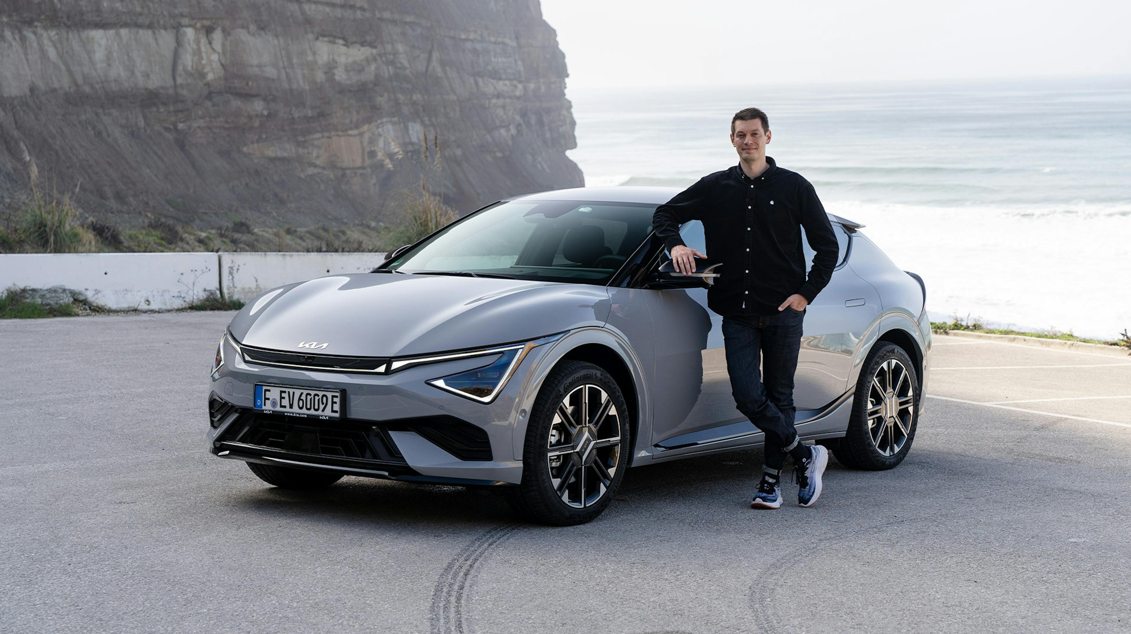 Grauer Kia EV6 Facelift mit einem Redakteur neben dem Auto, im Hintergrund das Meer.