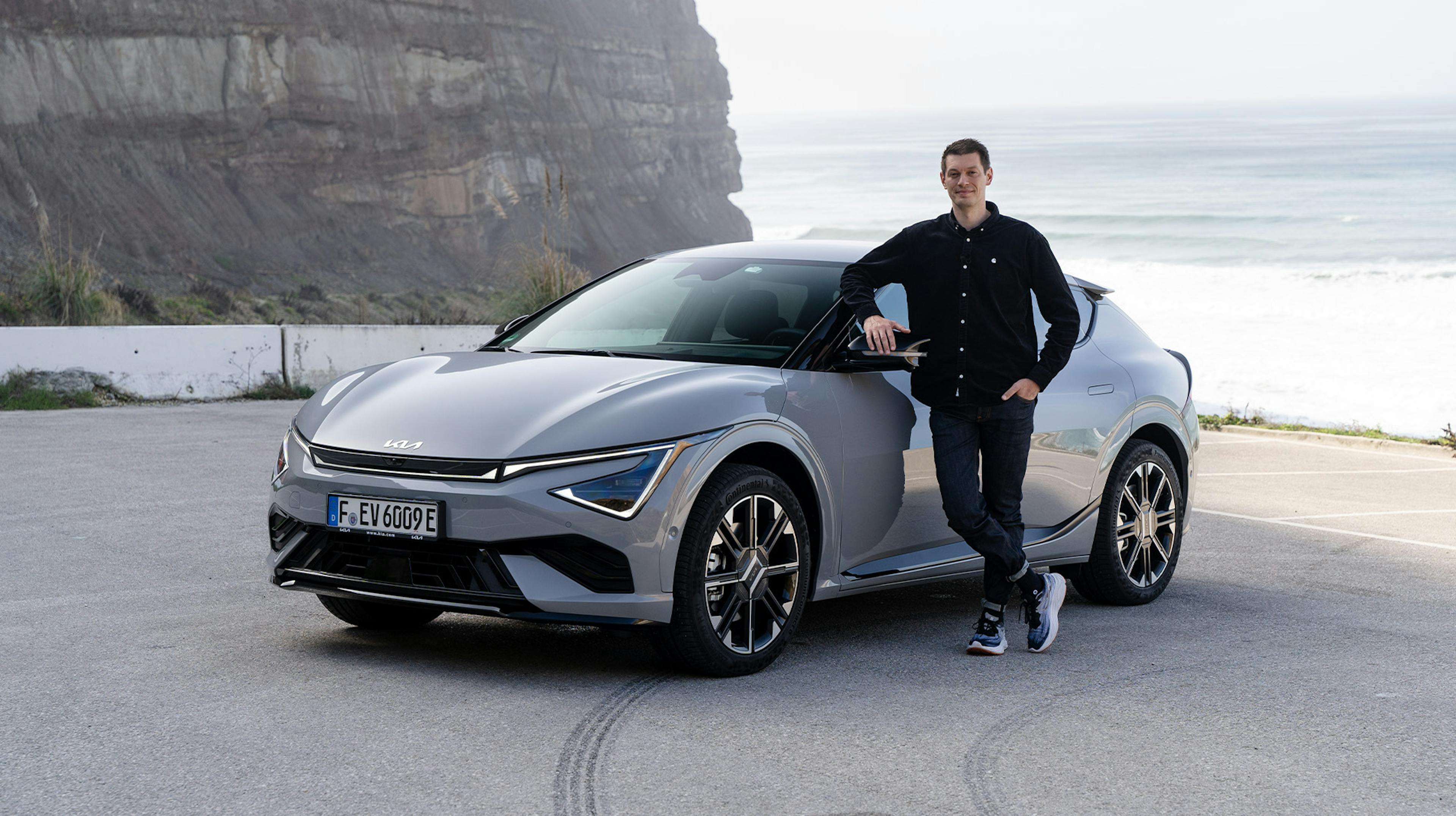  Grauer Kia EV6 Facelift mit einem Redakteur neben dem Auto, im Hintergrund das Meer.