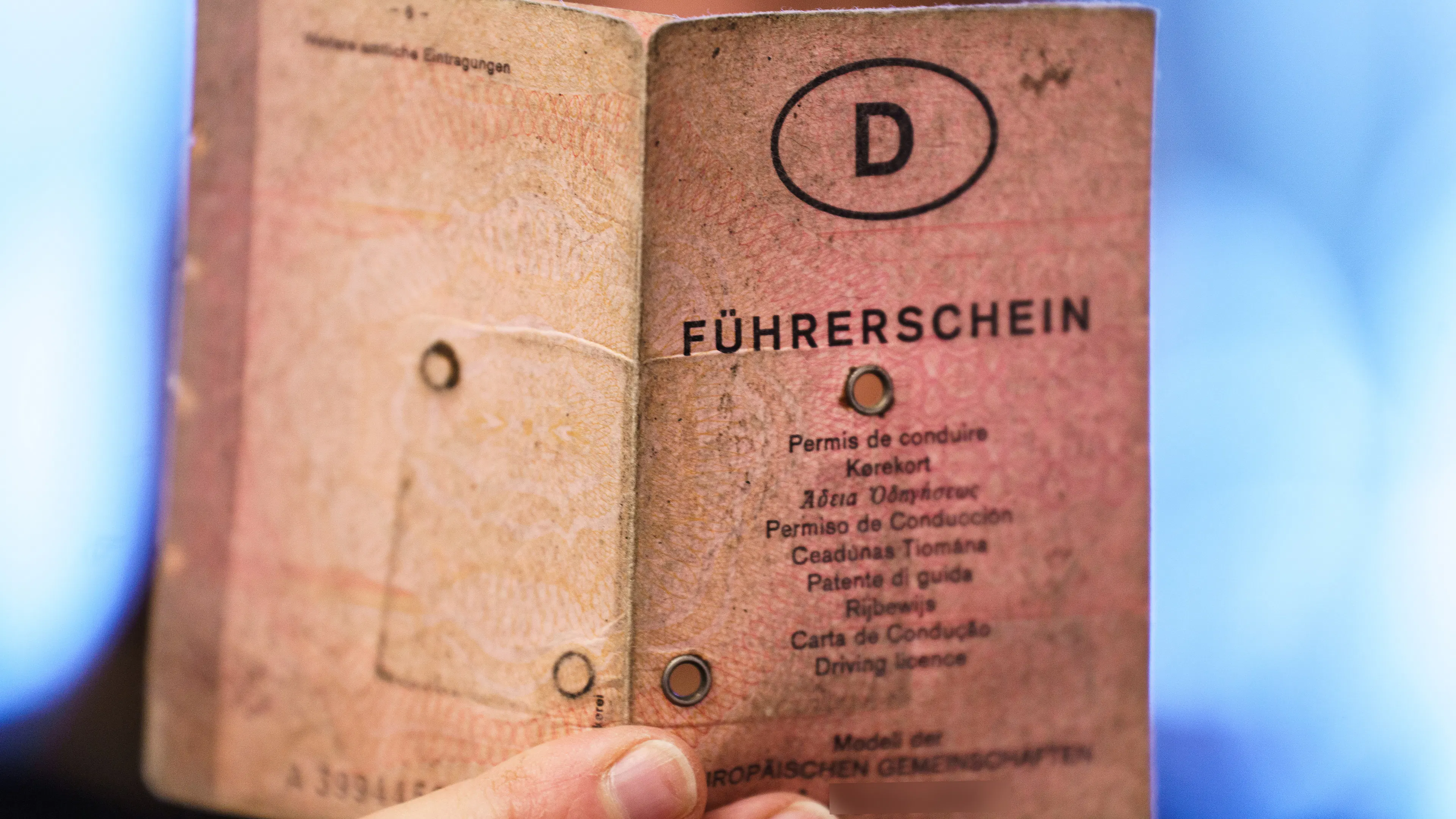 An old, pink German driver's license in paper format is held by one hand, with a capital "D" and the inscription "Führerschein" in different languages on it.