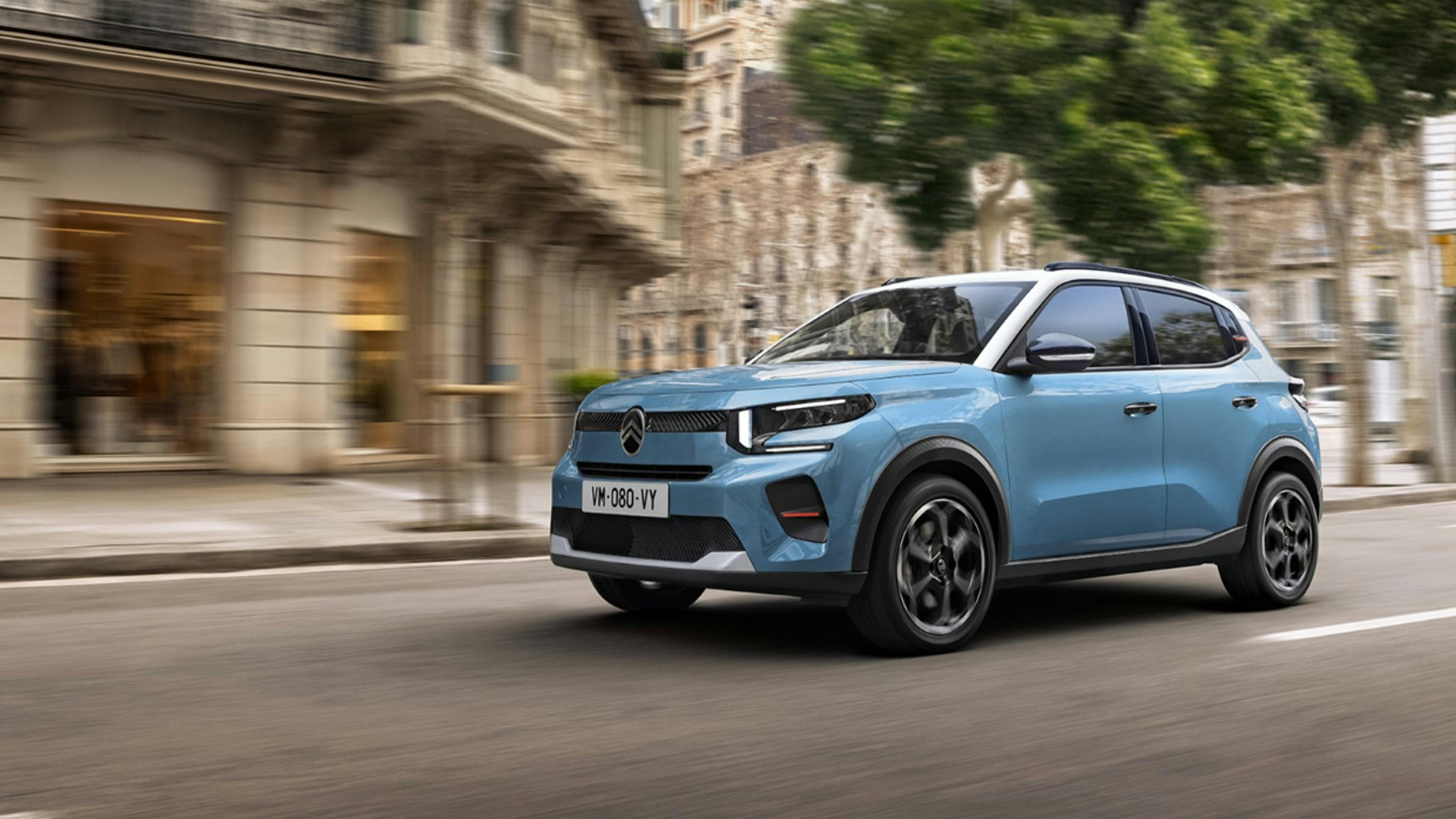 A blue Citroën ë-C3 drives from right to left in front of a façade.