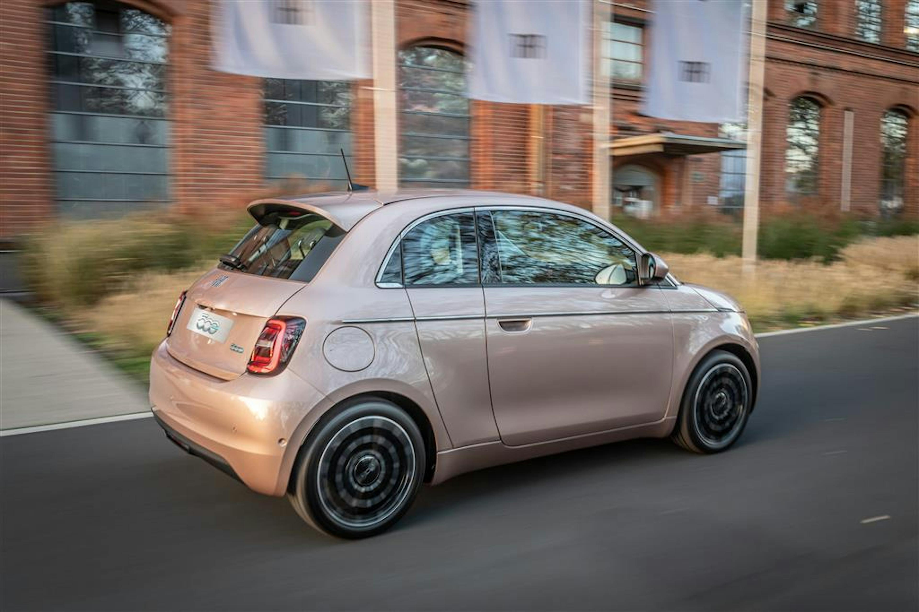 Ein roséfarbener Fiat 500 fährt von links nach rechts vor einer Industriefassade entlang.